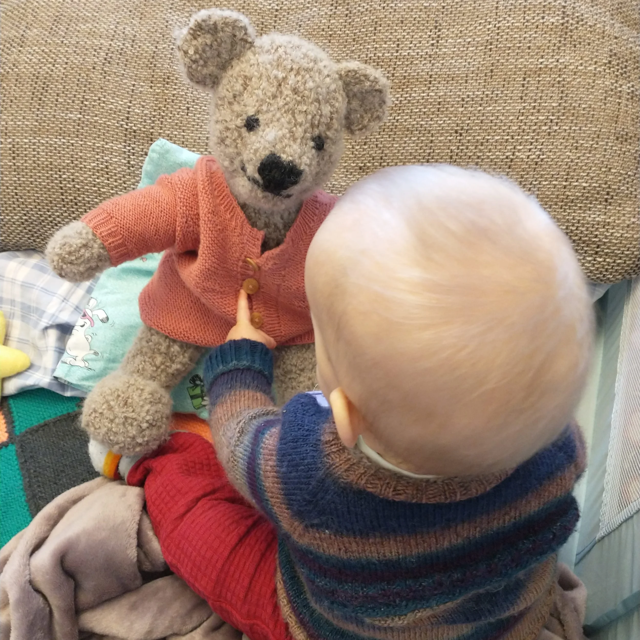 Ein Baby sitzt vor einem Plüschteddy, der einen roten Pullover trägt, während das Baby eine Hand auf den Bauch des Teddys legt.