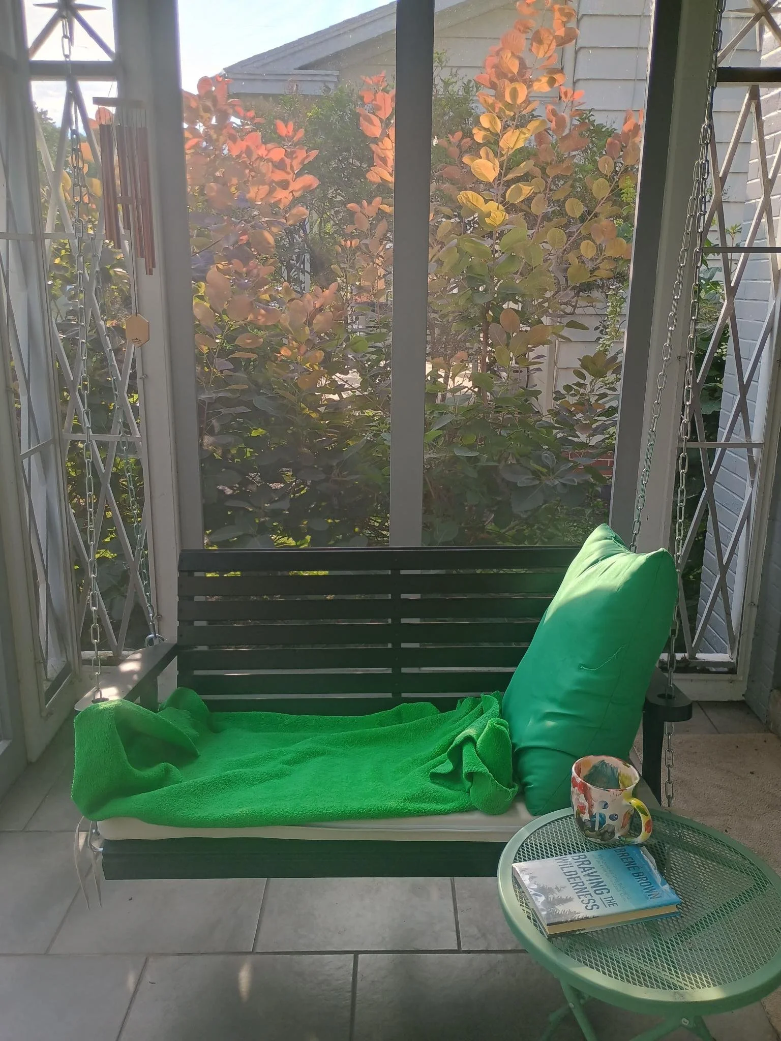 Photo of a porch swing with a green cushion, and a coffee mug and book rest next to it on a table.