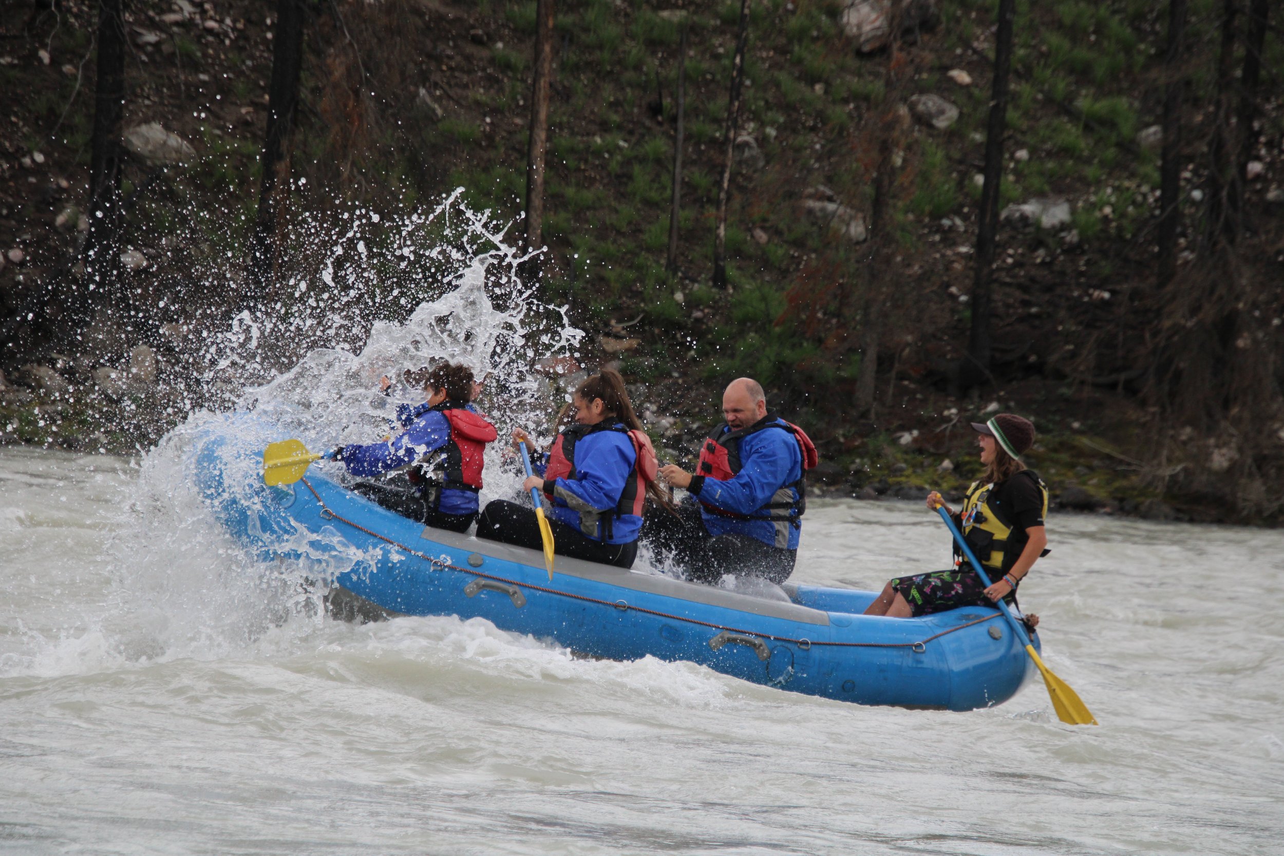 Four people white water rafting on a river with trees in the background. They are wearing life jackets and holding paddles.