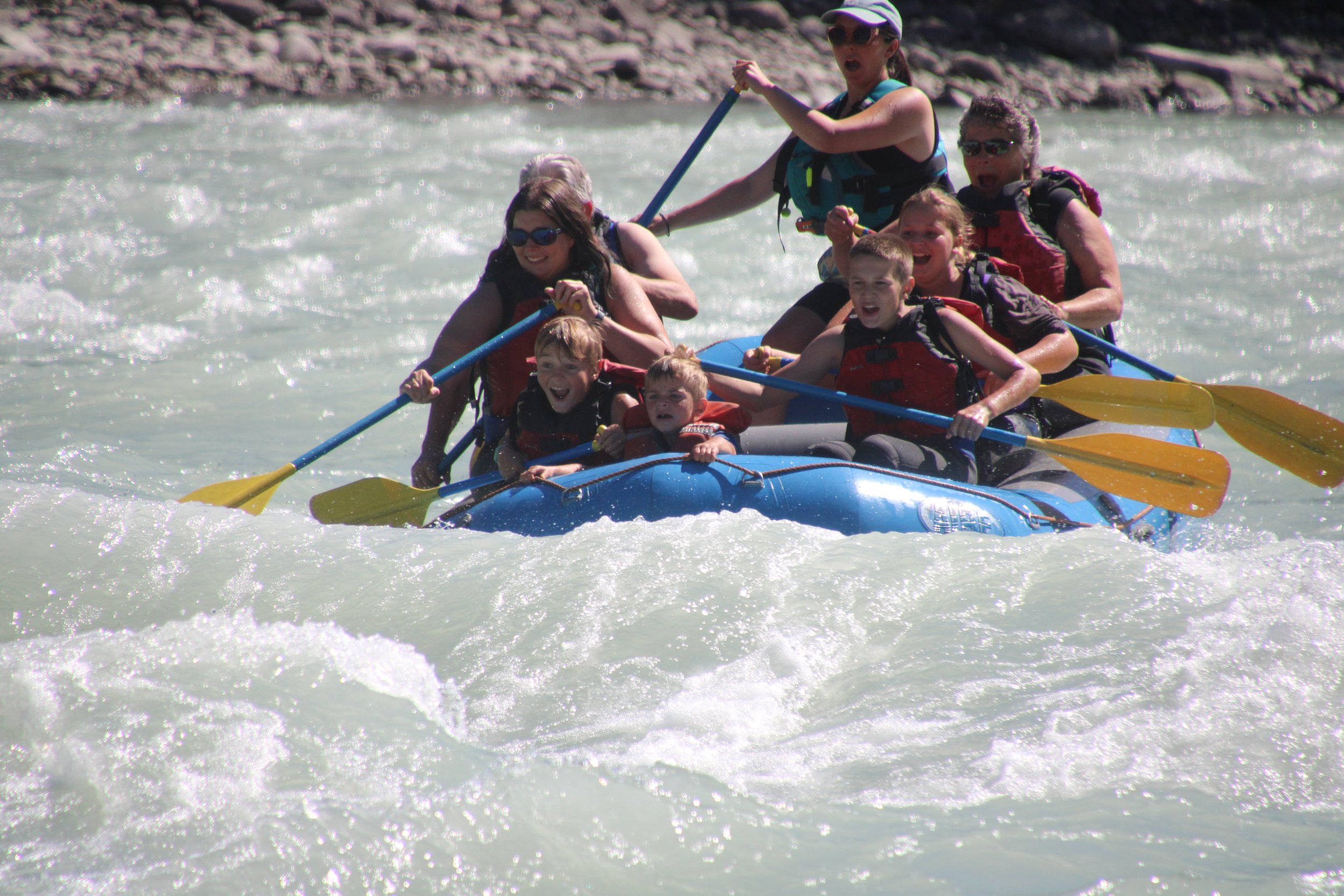 A group of kids and adults white water rafting on a river. They are wearing life jackets and holding yellow paddles, enjoying the adventure on a sunny day.