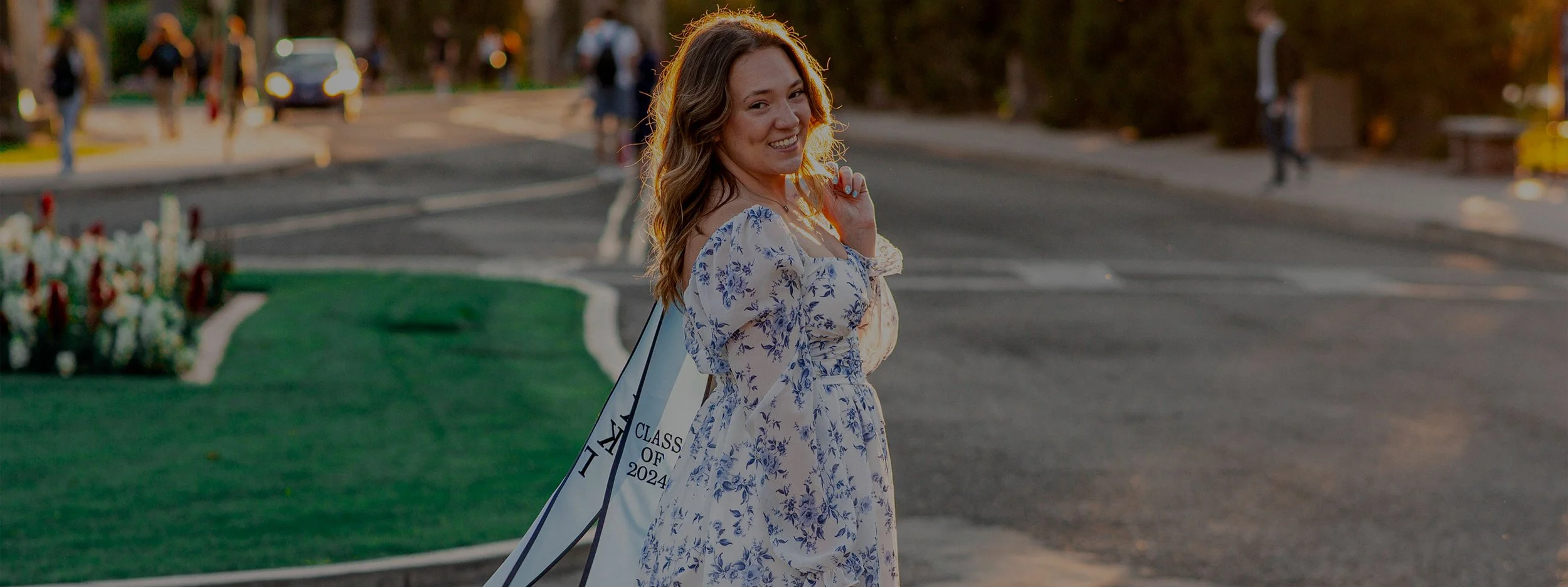A young woman in a floral dress smiling and walking outside during sunset, holding a sash that says 'Class of 2024'.