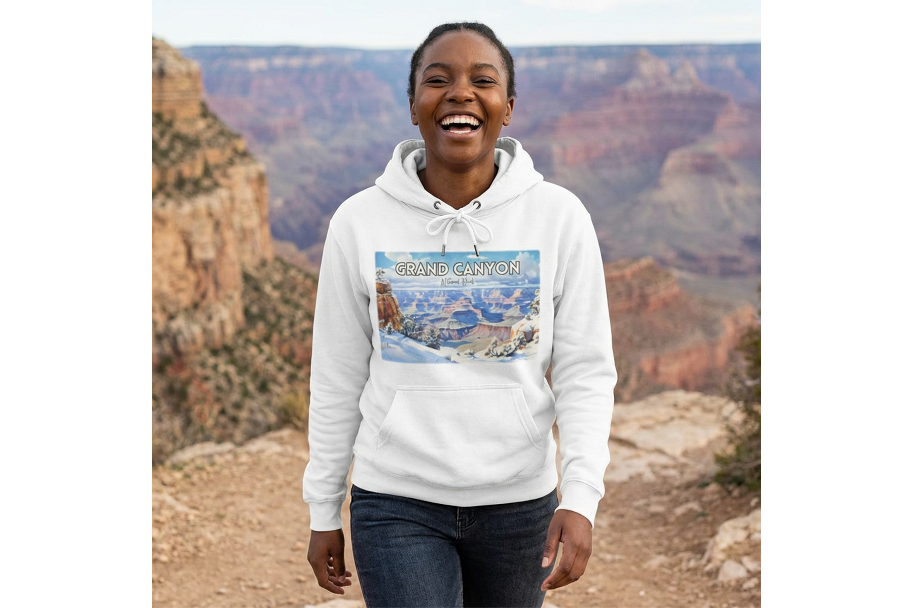 A woman smiling at the camera during a visit to the Grand Canyon, wearing a white hoodie with a Grand Canyon graphic and trees in the background.