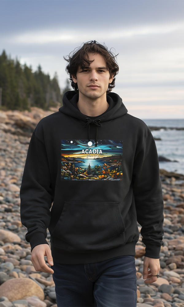 Model wearing a black hoodie with colorful Acadia National Park landscape artwork, standing on a rocky shoreline with ocean water and evergreen trees in the background.