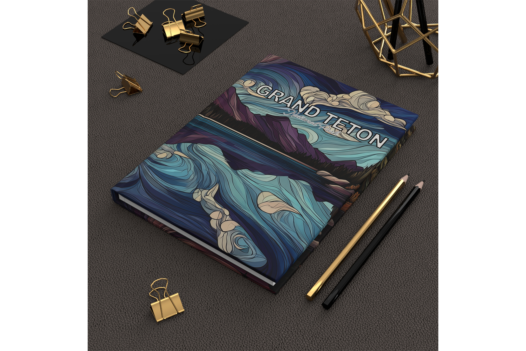 Grand Teton art journal with swirling blue mountain landscape cover, shown on a desk with pencils and gold binder clips.