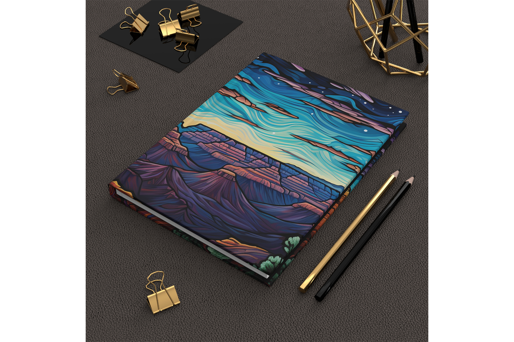 A closed notebook with a colorful, abstract landscape cover featuring mountains, sky, and water, placed on a black textured surface. Next to it are two pencils and two gold binder clips, with additional gold binder clips and a geometric gold desk organizer visible nearby.