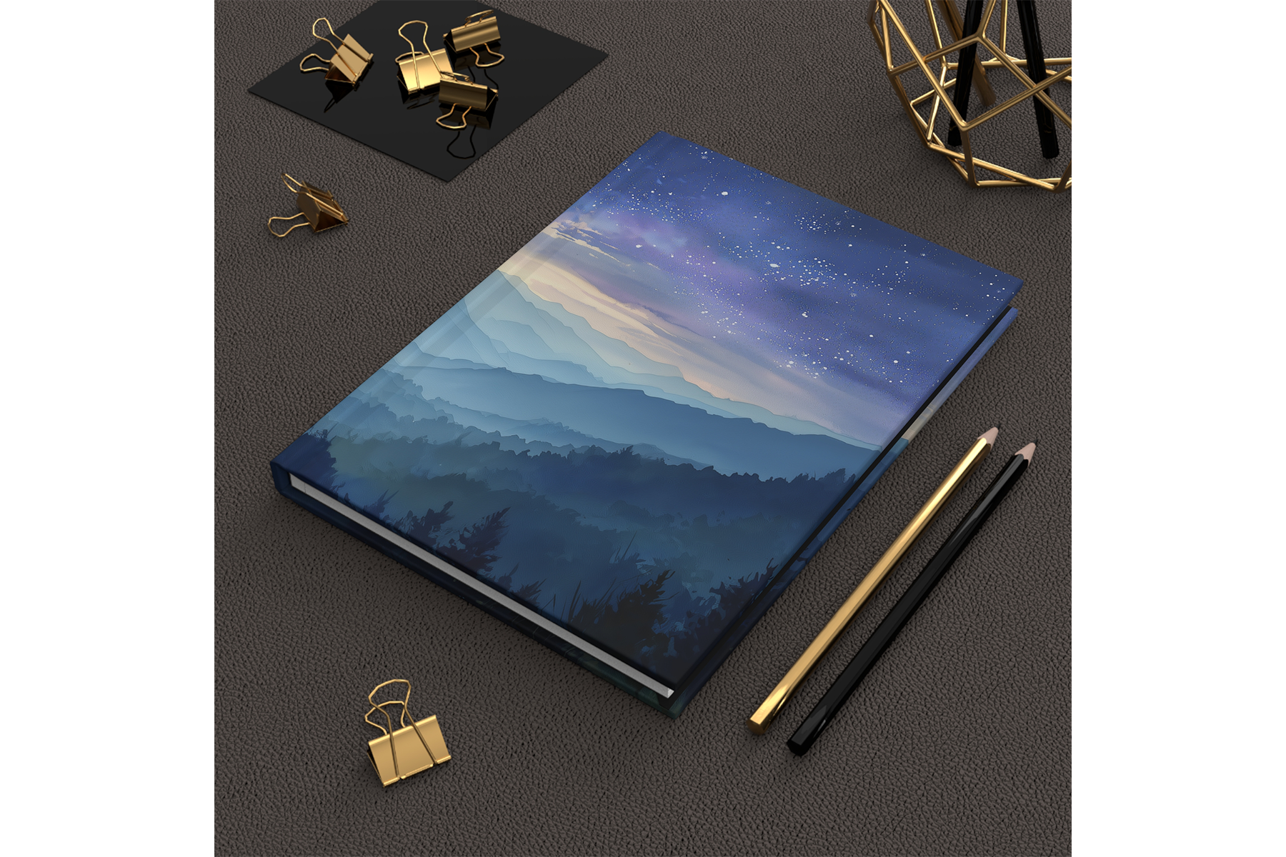 A closed hardcover book with a mountain landscape and starry sky illustration on the cover, placed on a dark textured surface. Nearby are gold and black pens, and gold binder clips are scattered around.