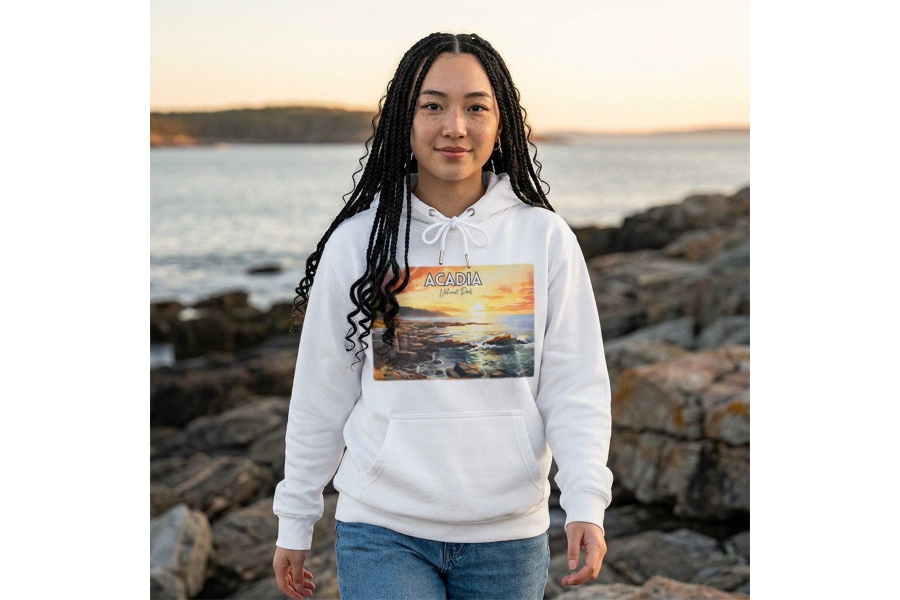 Woman wearing a white Acadia National Park hoodie with sunset coastal artwork, standing on a rocky shoreline by the water.