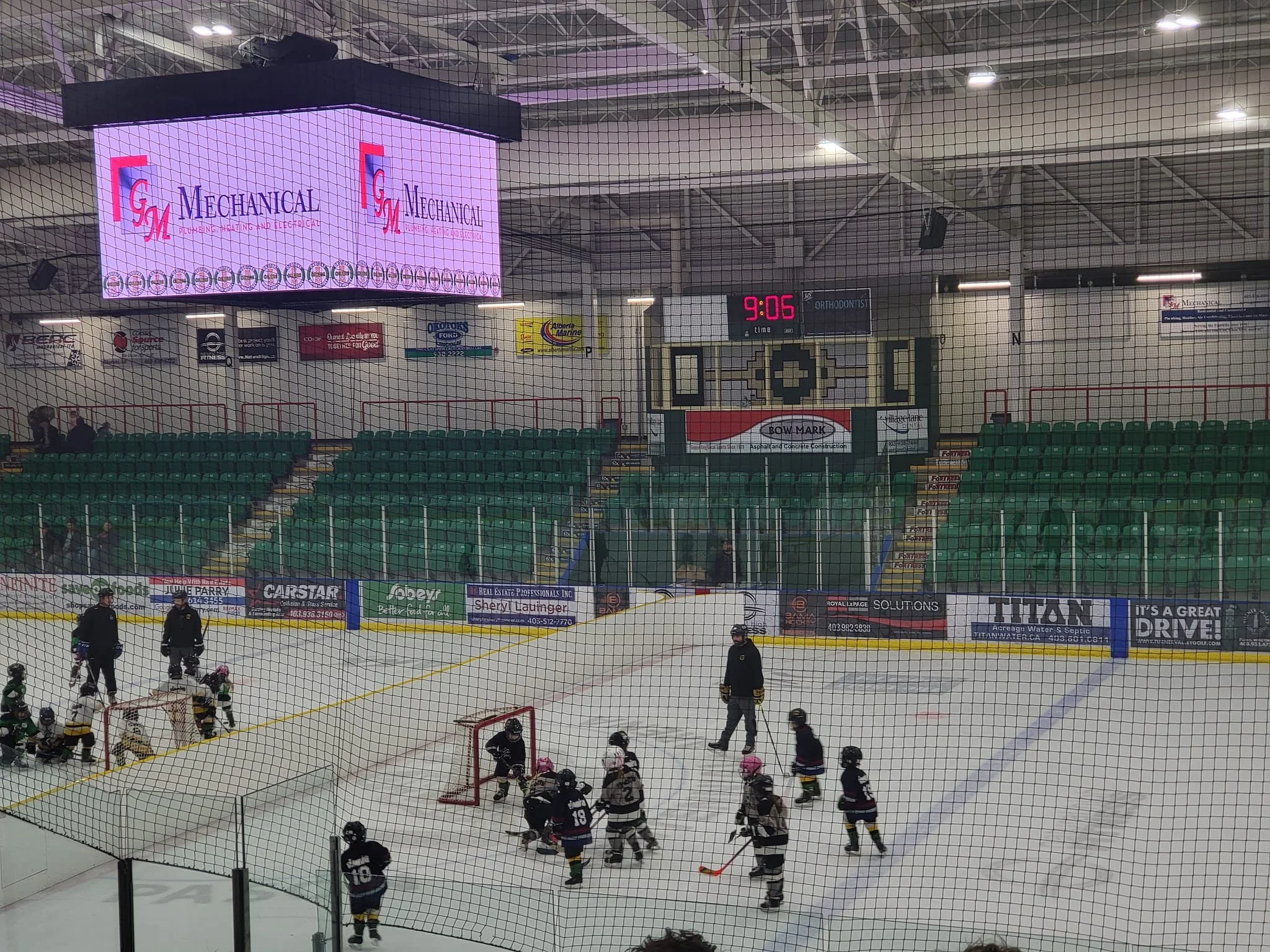 Indoor ice hockey rink with young players, coaches, and parents on the ice and in the stands, digital scoreboard showing 9:06, and large electronic display board.