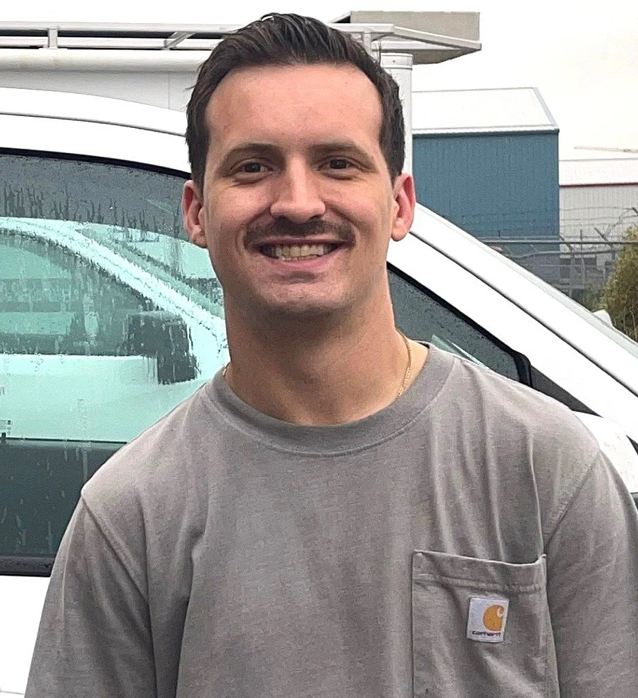 A smiling man with dark hair wearing a gray Carhartt t-shirt, standing outdoors in front of a white vehicle with a wet windshield and a building in the background.
