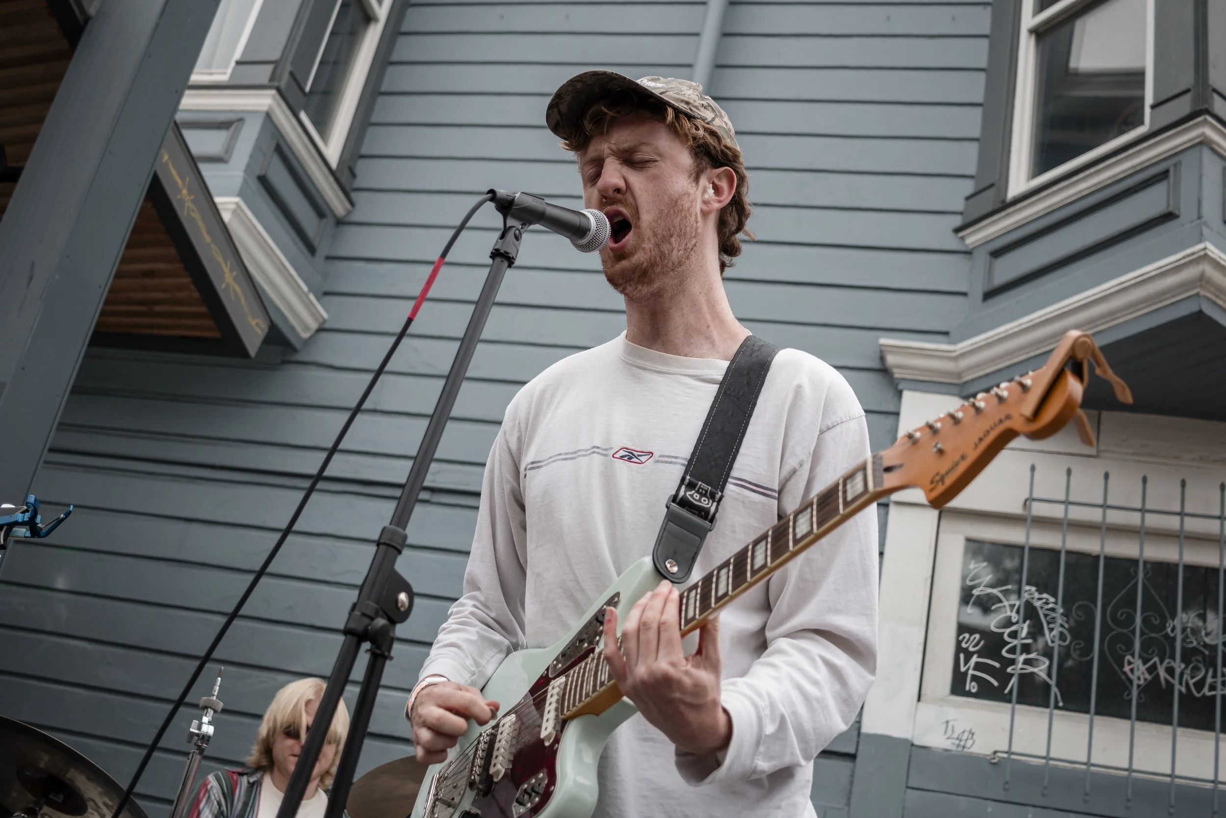A man singing into a microphone while playing an electric guitar outdoors, with a woman playing drums in the background, in front of a blue house with windows.