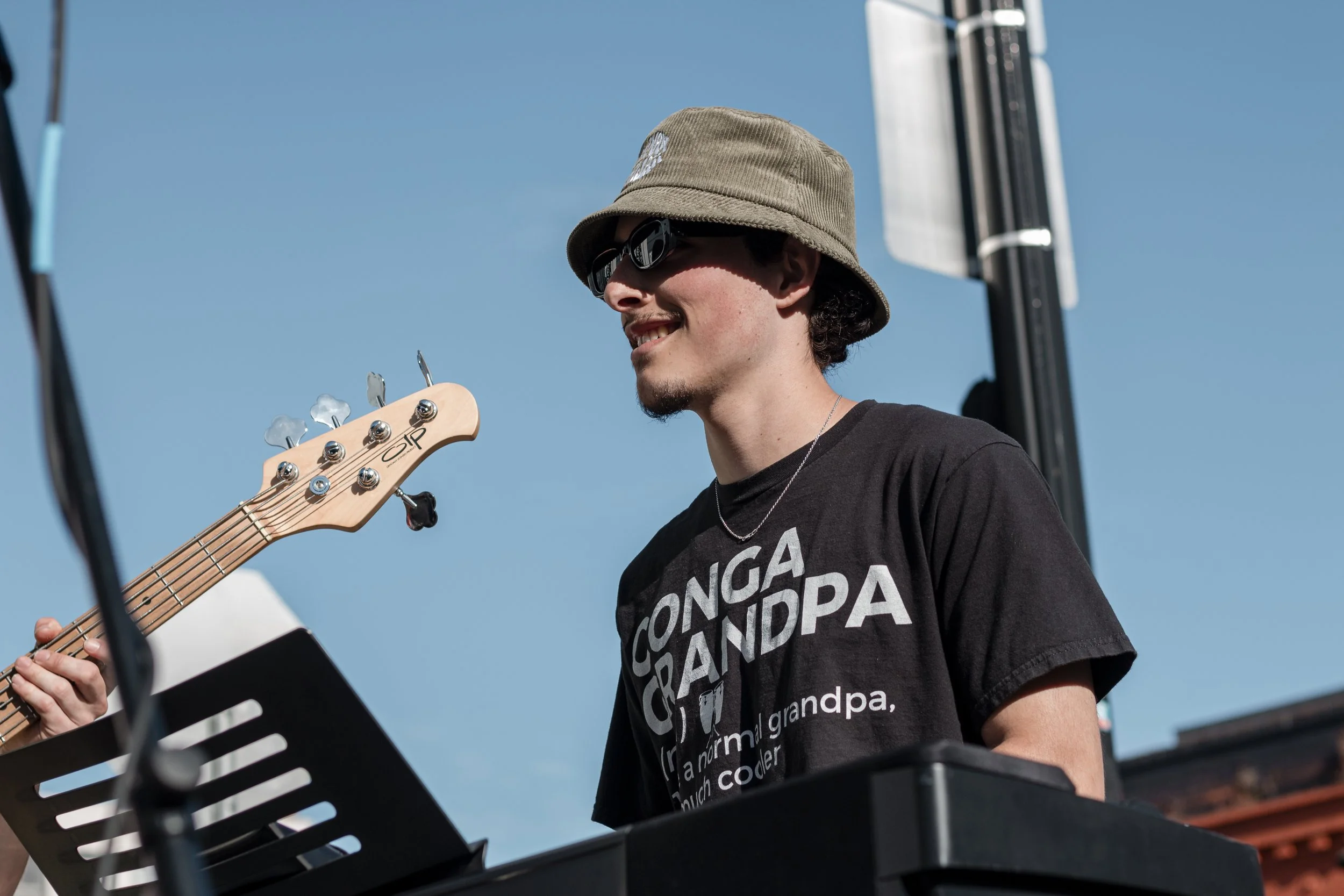 A young man wearing sunglasses, a bucket hat, and a black t-shirt with the text 'CONGA OR ANDPA' is smiling while playing a keyboard during an outdoor event against a blue sky.