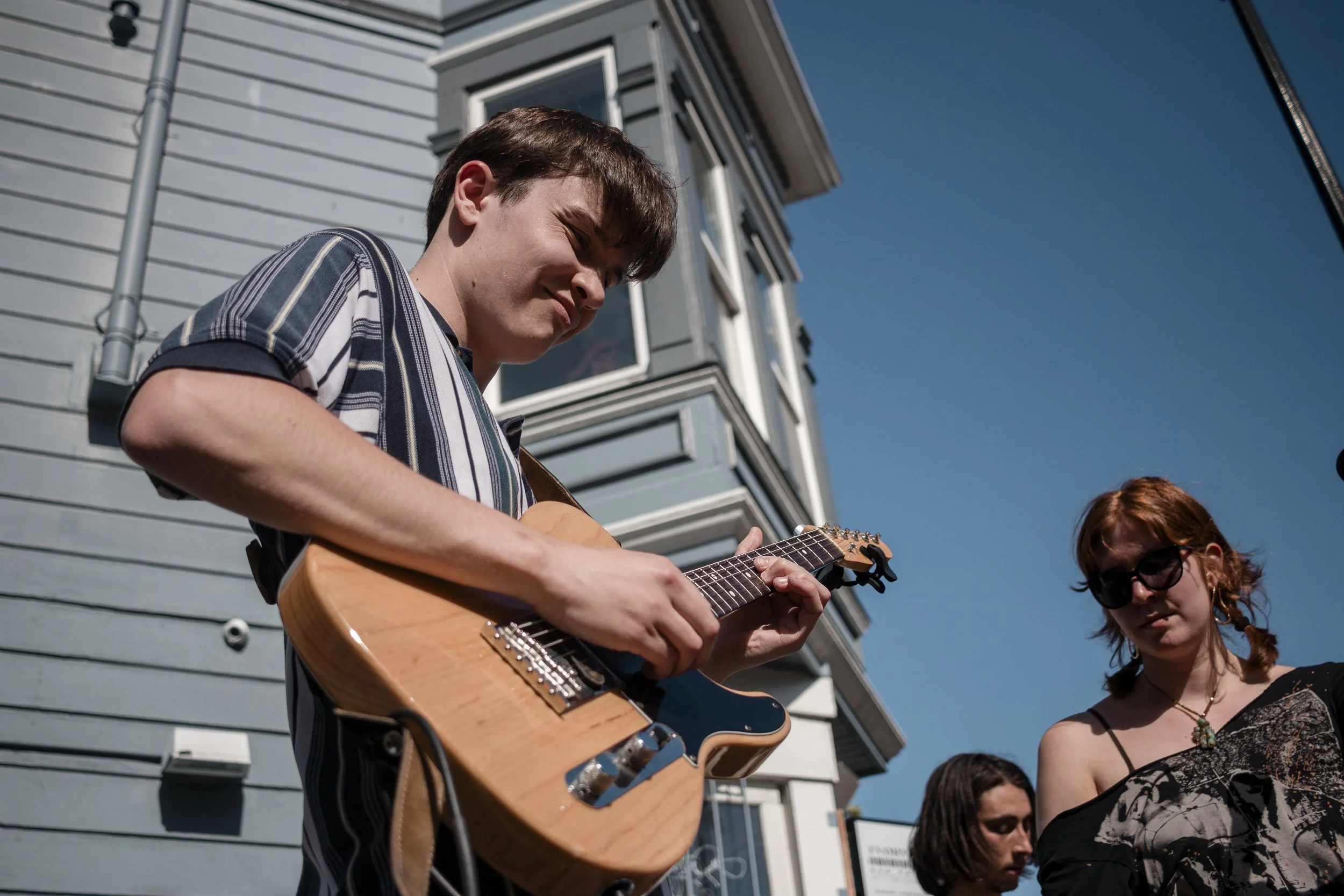 A young man playing an acoustic guitar outdoors with a blue sky in the background, next to a light blue house. Two women and a man are nearby, one woman wearing sunglasses and a black patterned top.