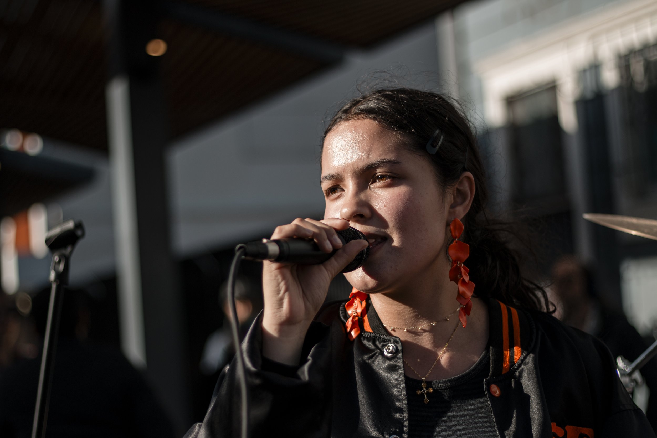 A woman singing into a microphone during an outdoor performance, wearing orange earrings and a black jacket with orange accents.