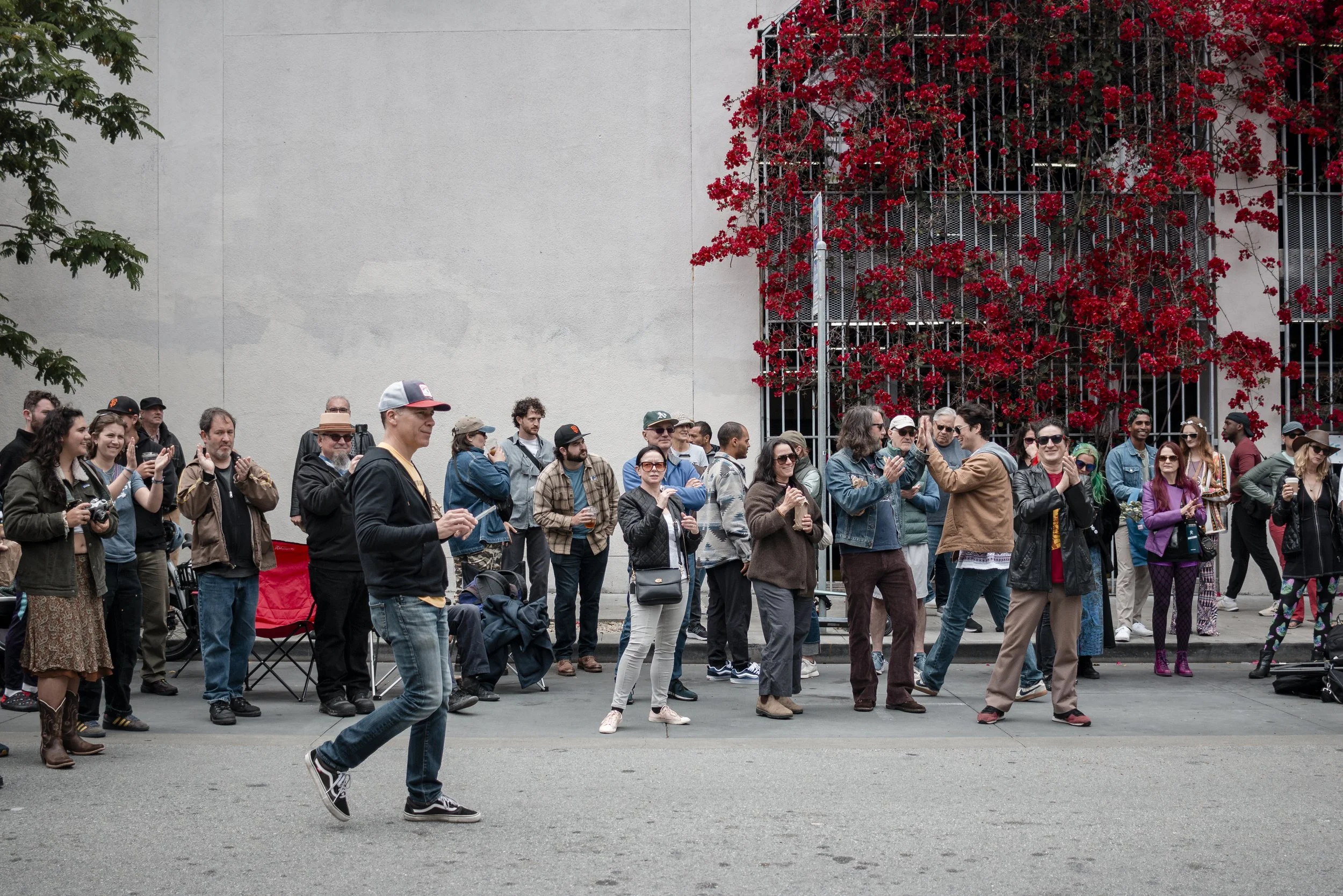 Group of people standing and watching a performer on the street during daytime, with a building wall and red flowering vine in the background.