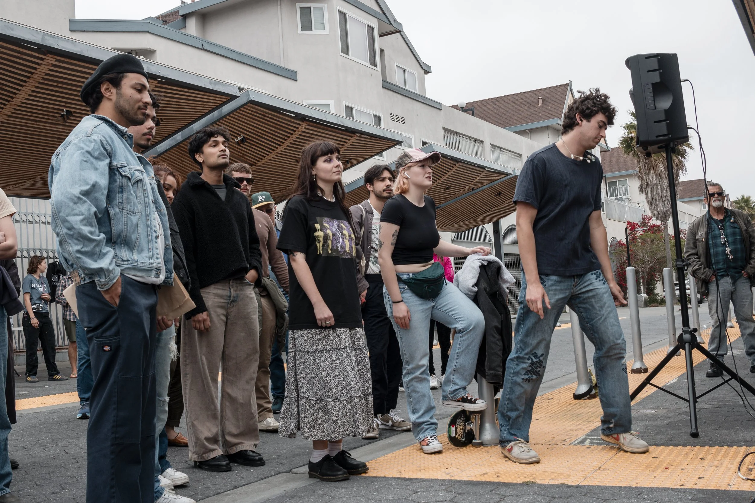 A group of people standing outdoors in front of a building, some with their hands in their pockets, others looking down or to the side, with a speaker and a man in sunglasses on the right.