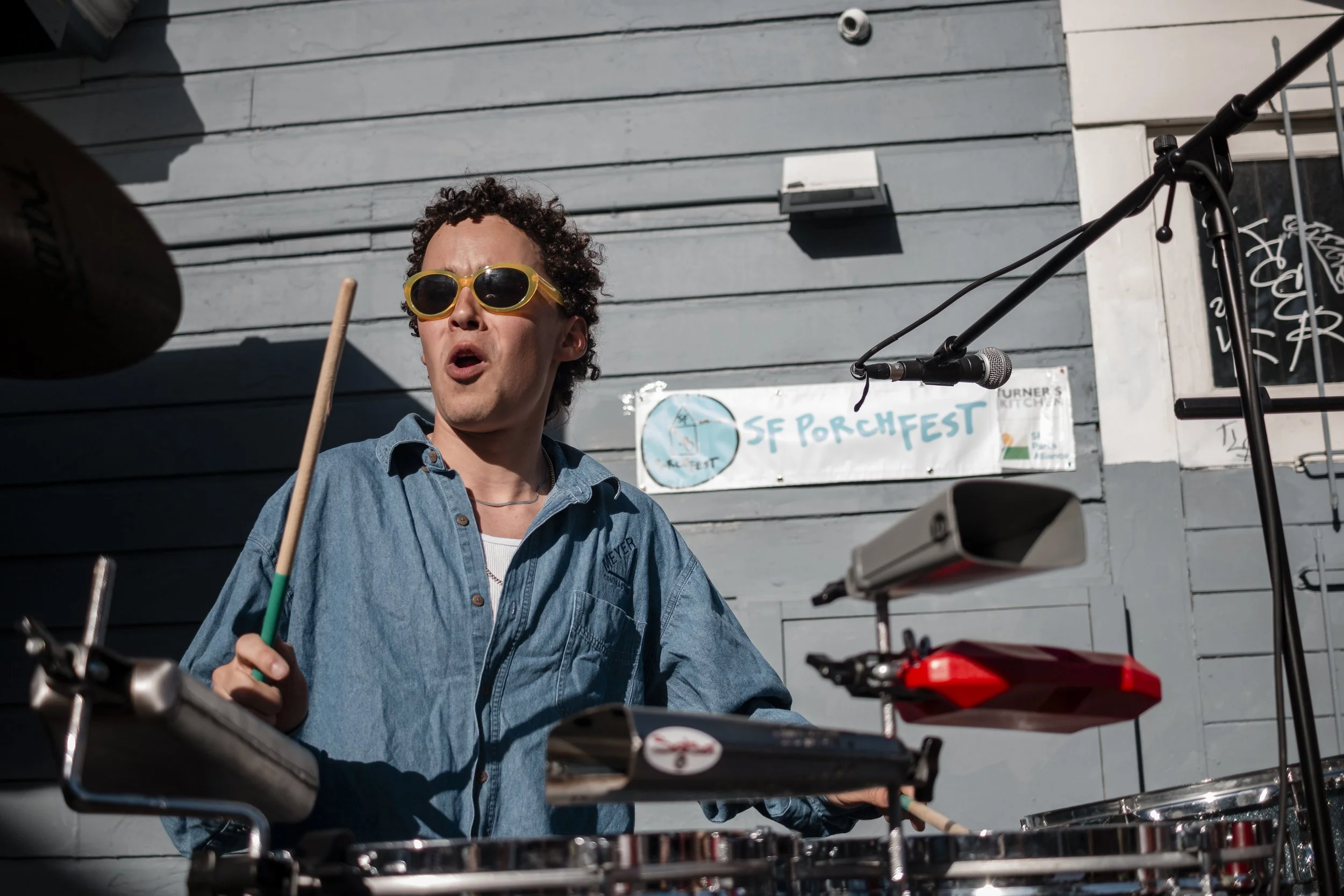 Person playing drums and singing into a microphone at an outdoor event, wearing sunglasses and a denim shirt, with a sign that reads 'SF Porchfest' in the background.