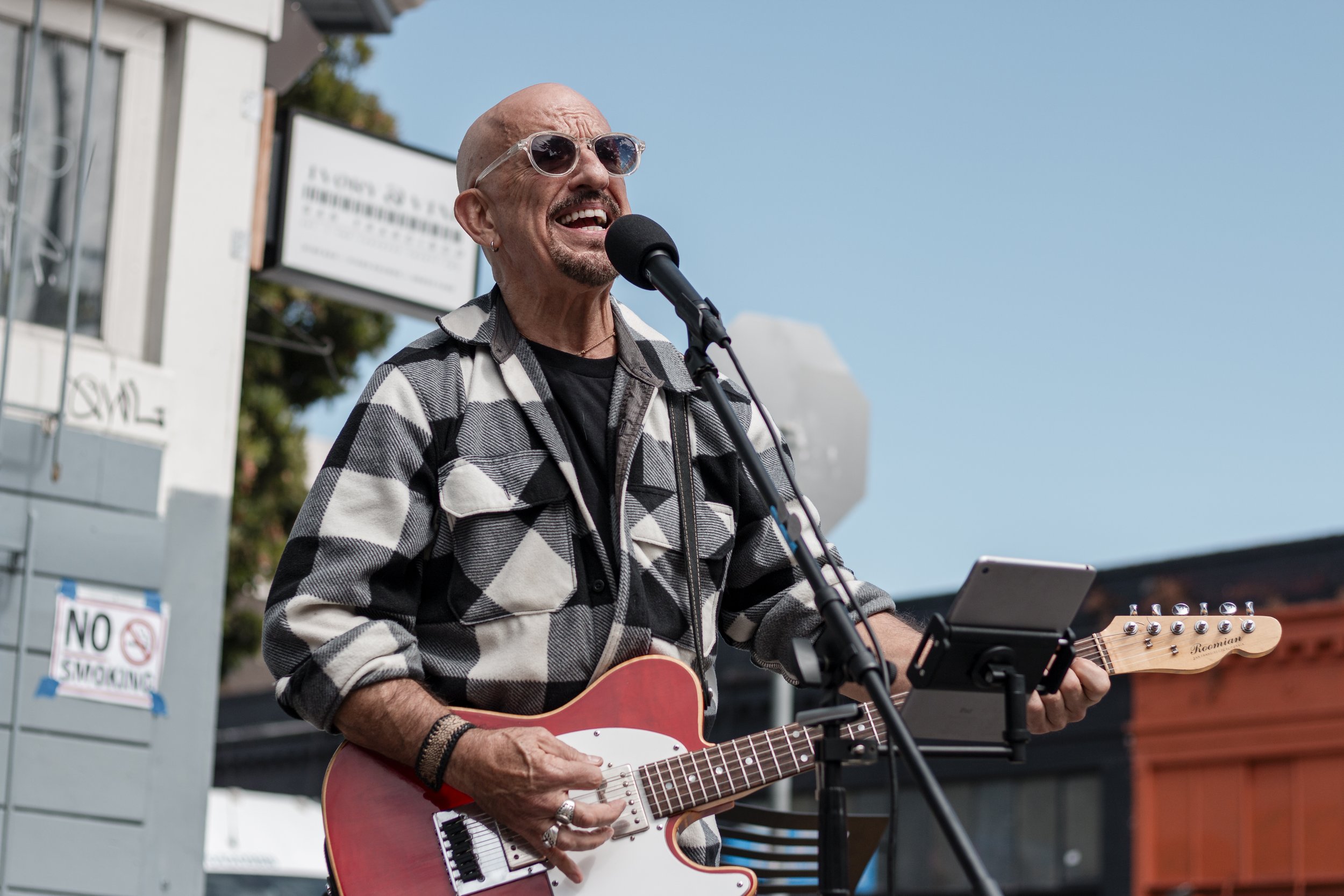 A man with sunglasses and a black and white checkered jacket singing into a microphone while playing an electric guitar outdoors on a sunny day.