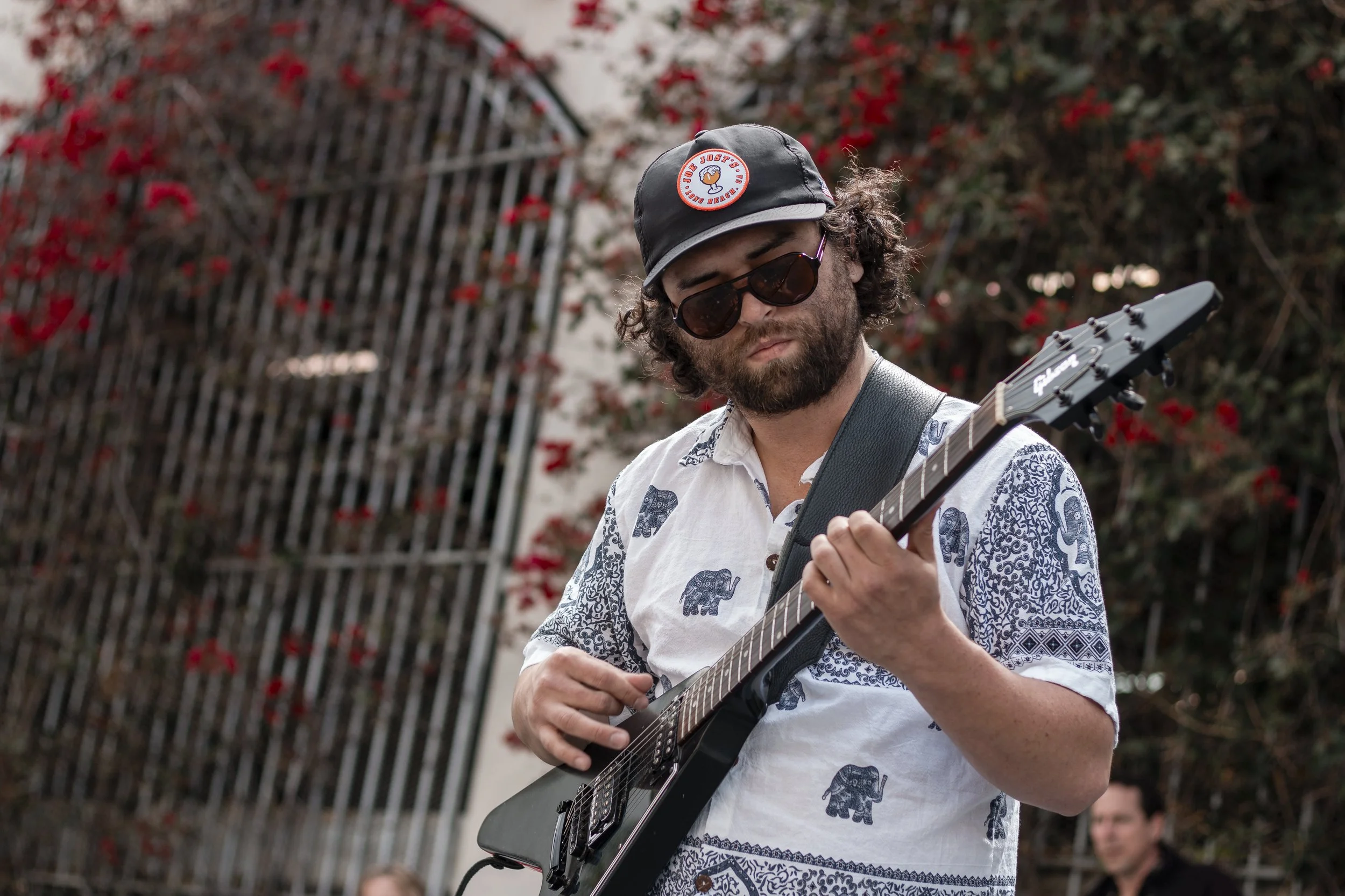 A man with curly hair and a beard playing an electric guitar outdoors. He is wearing dark sunglasses, a black cap with a circular logo, and a white shirt with blue elephant patterns. In the background, there is a metal gate and some red flowering pla