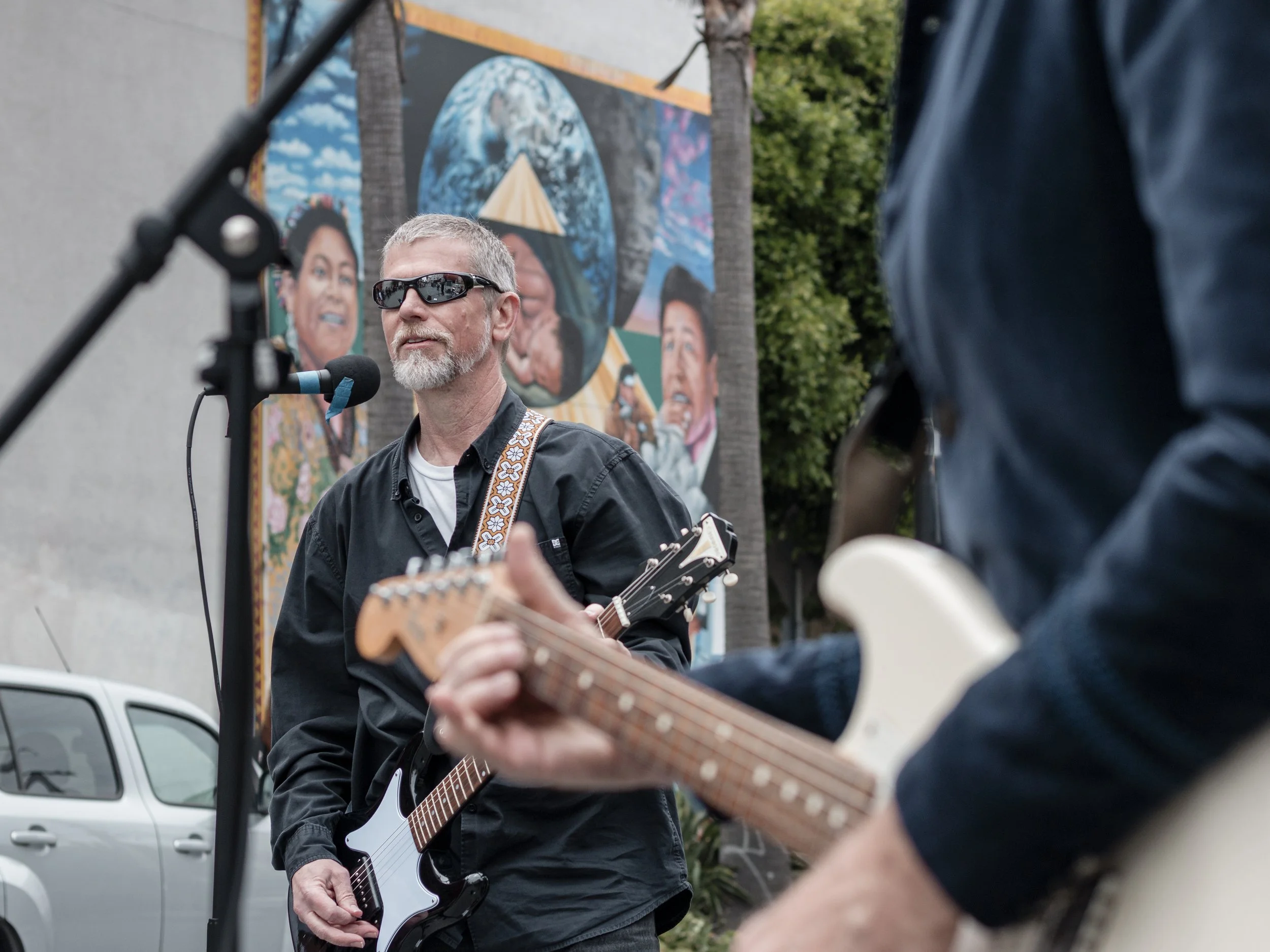 A man with gray hair, beard, and sunglasses playing a guitar and singing into a microphone outdoors. A mural with faces and abstract art is visible in the background, along with some trees and a parked car.