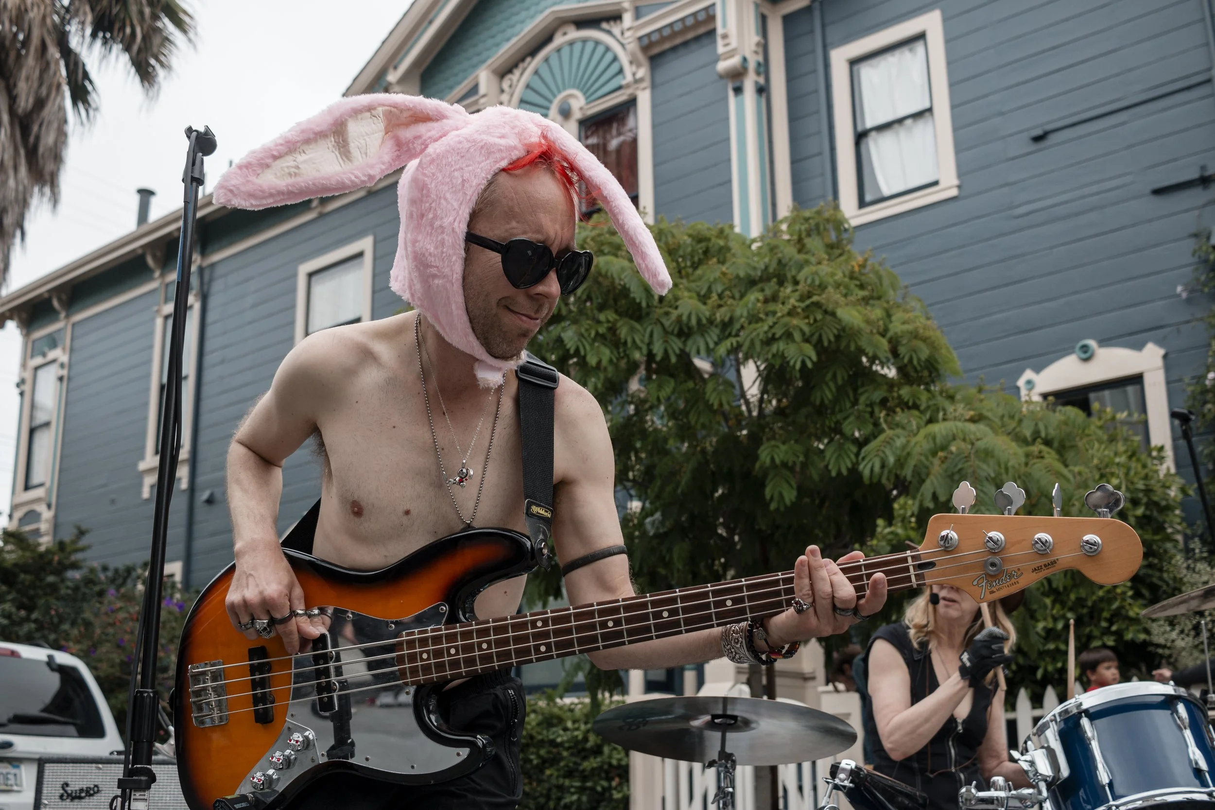 A shirtless man wearing sunglasses and a pink bunny hat playing an electric bass guitar outdoors, with a woman playing drums in the background.
