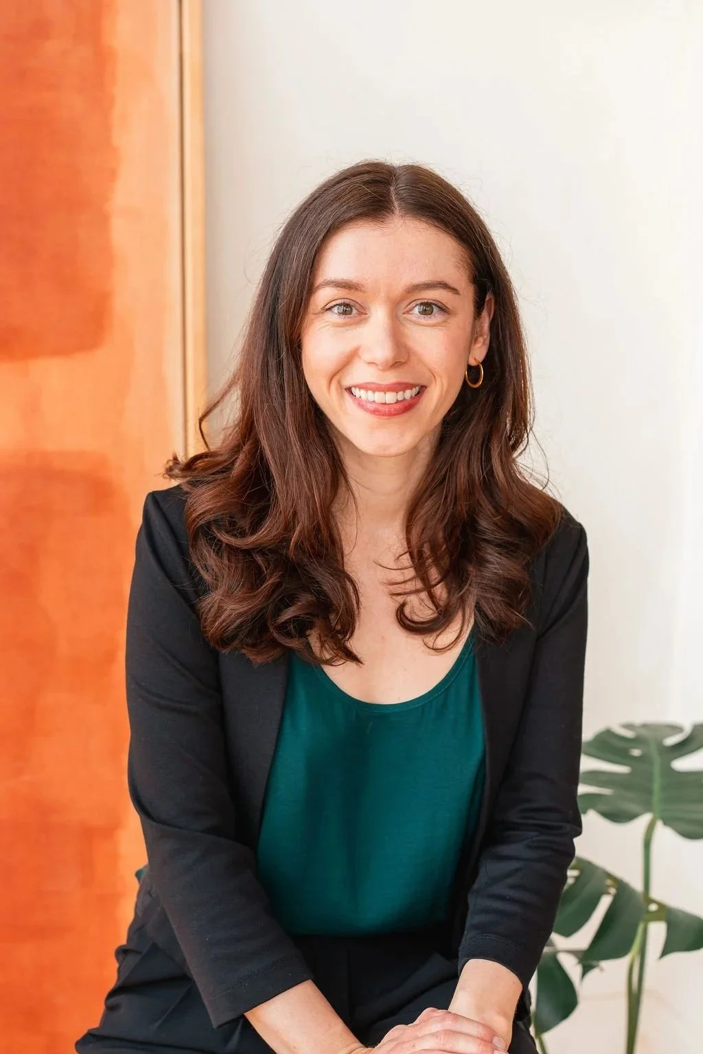 Mira Dineen is smiling confidently and wearing a black blazer and a teal top. She has shoulder length brown hair and is sitting indoors with a wooden and white background, and a green leafy plant nearby.
