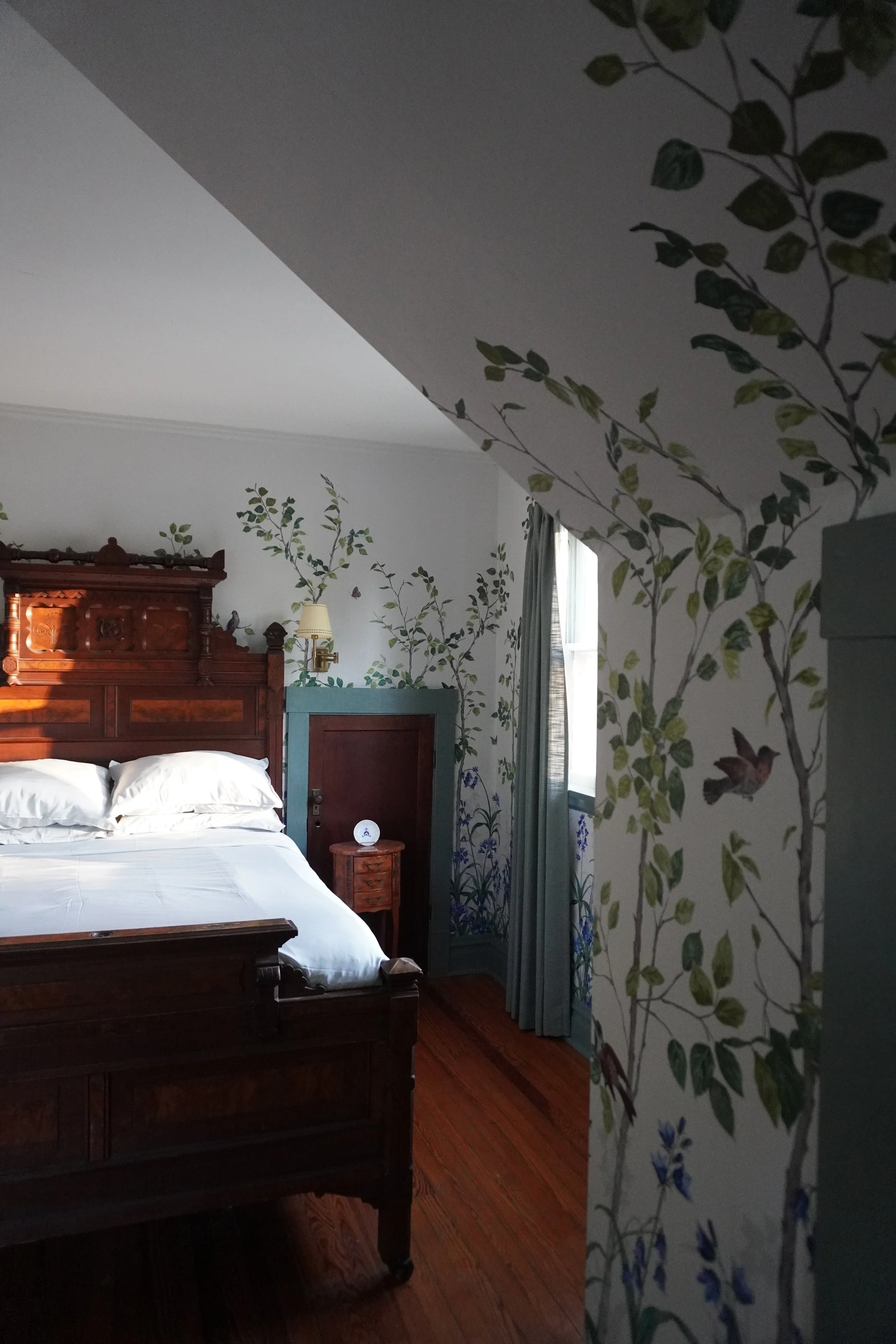 A cozy bedroom with a large wooden bed, a nightstand, and walls decorated with green leafy tree and bird mural, with a window and curtains in the background.