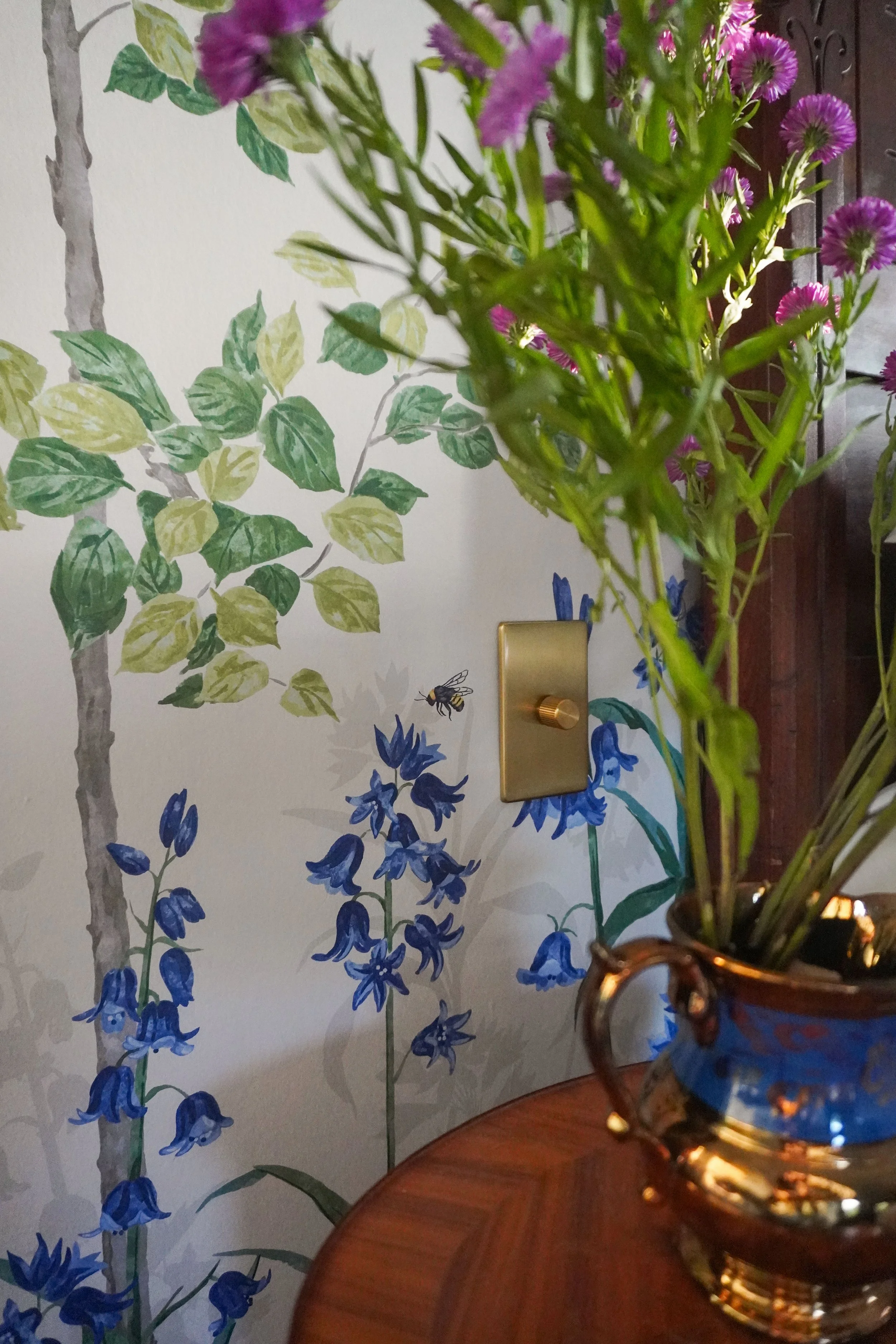 A decorative wall with painted green and yellow leaves, blue and purple flowers, and a bee, along with a brass light switch and part of a wooden table with a glass vase containing green and pink flowers.