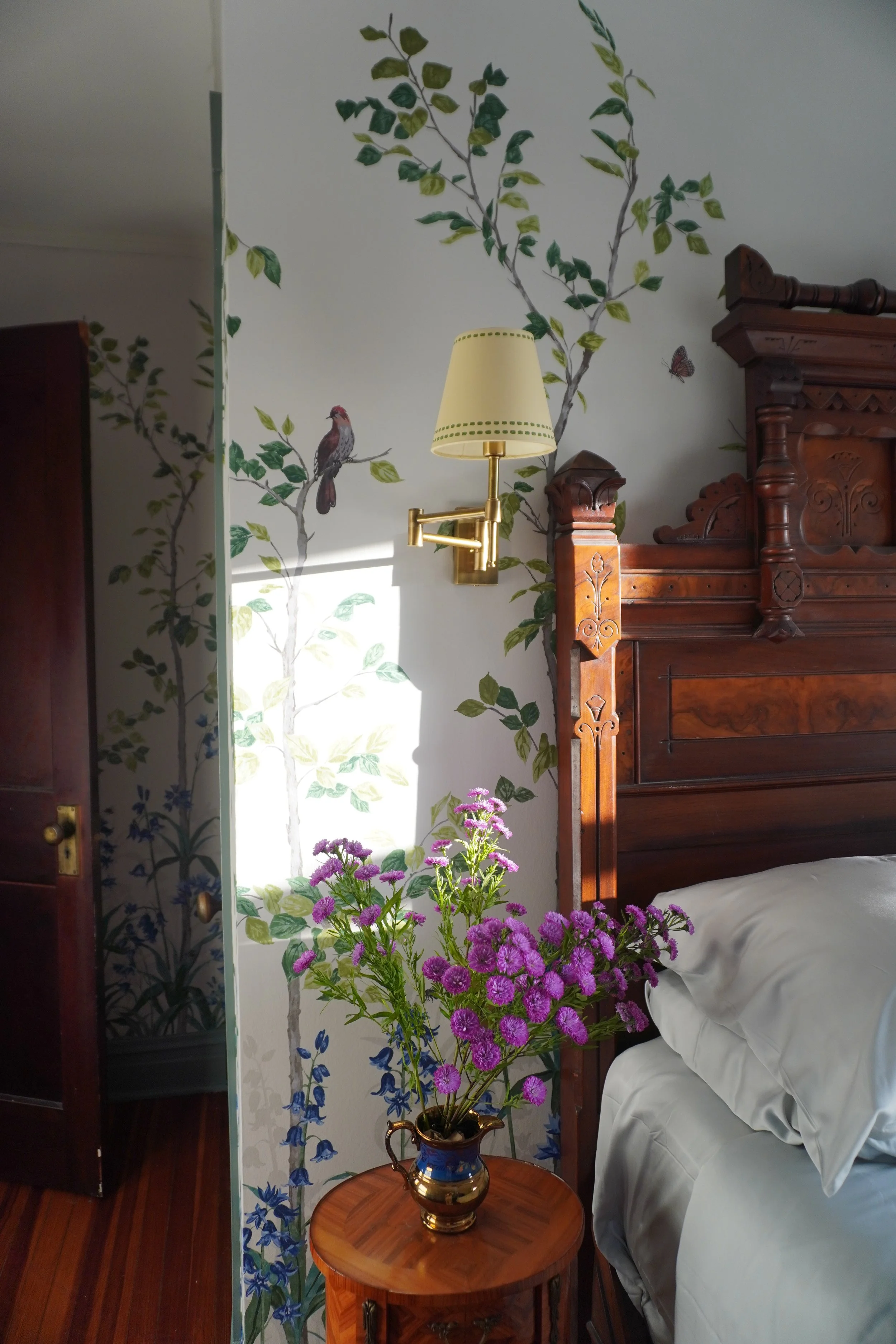 A bedroom corner with a wooden headboard, a bedside table with a vase of pink and purple flowers, a wall decorated with a nature mural featuring birds and leaves, a wall-mounted lamp, and part of a bed with white bedding.