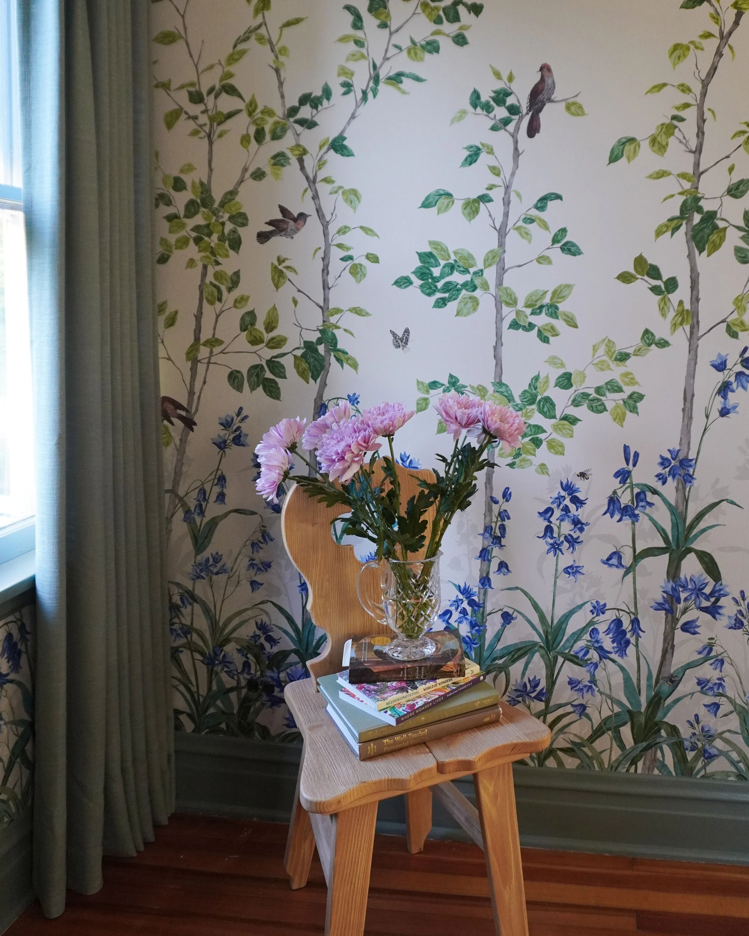 A wooden side table with a glass vase filled with pink and purple flowers, placed on top of a stack of books, next to a chair in front of a floral and bird-themed wallpaper.