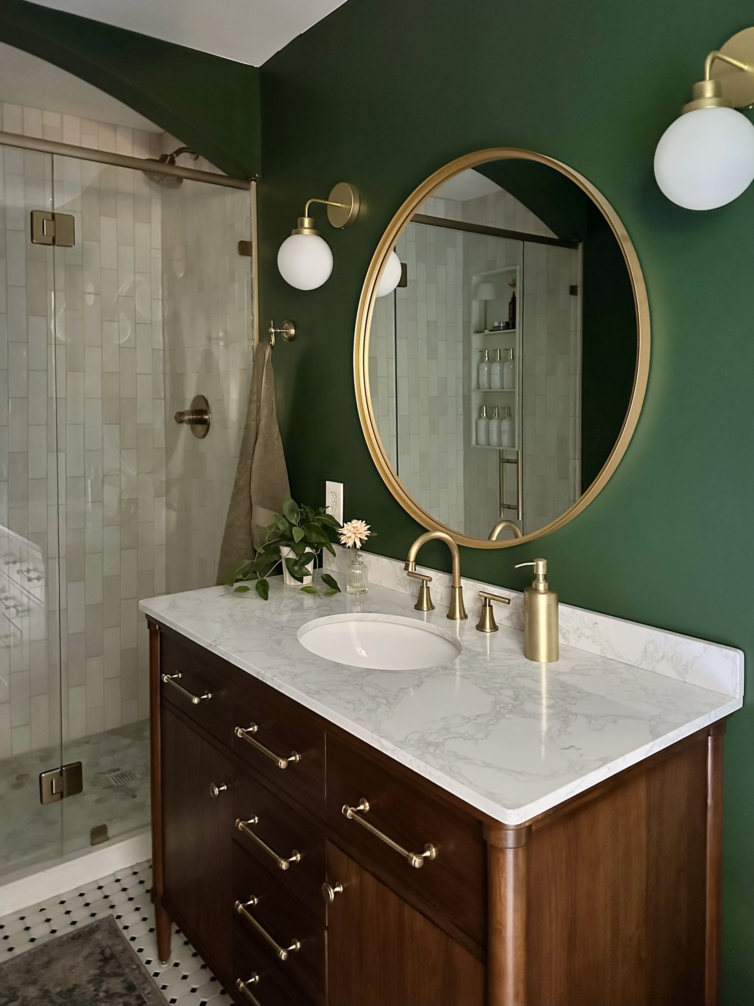A bathroom vanity with a dark wood cabinet, white marble countertop, undermount sink, and brass hardware. Above the sink is an oval mirror with a gold frame, flanked by two wall-mounted white globe light fixtures with brass accents. There is a small 