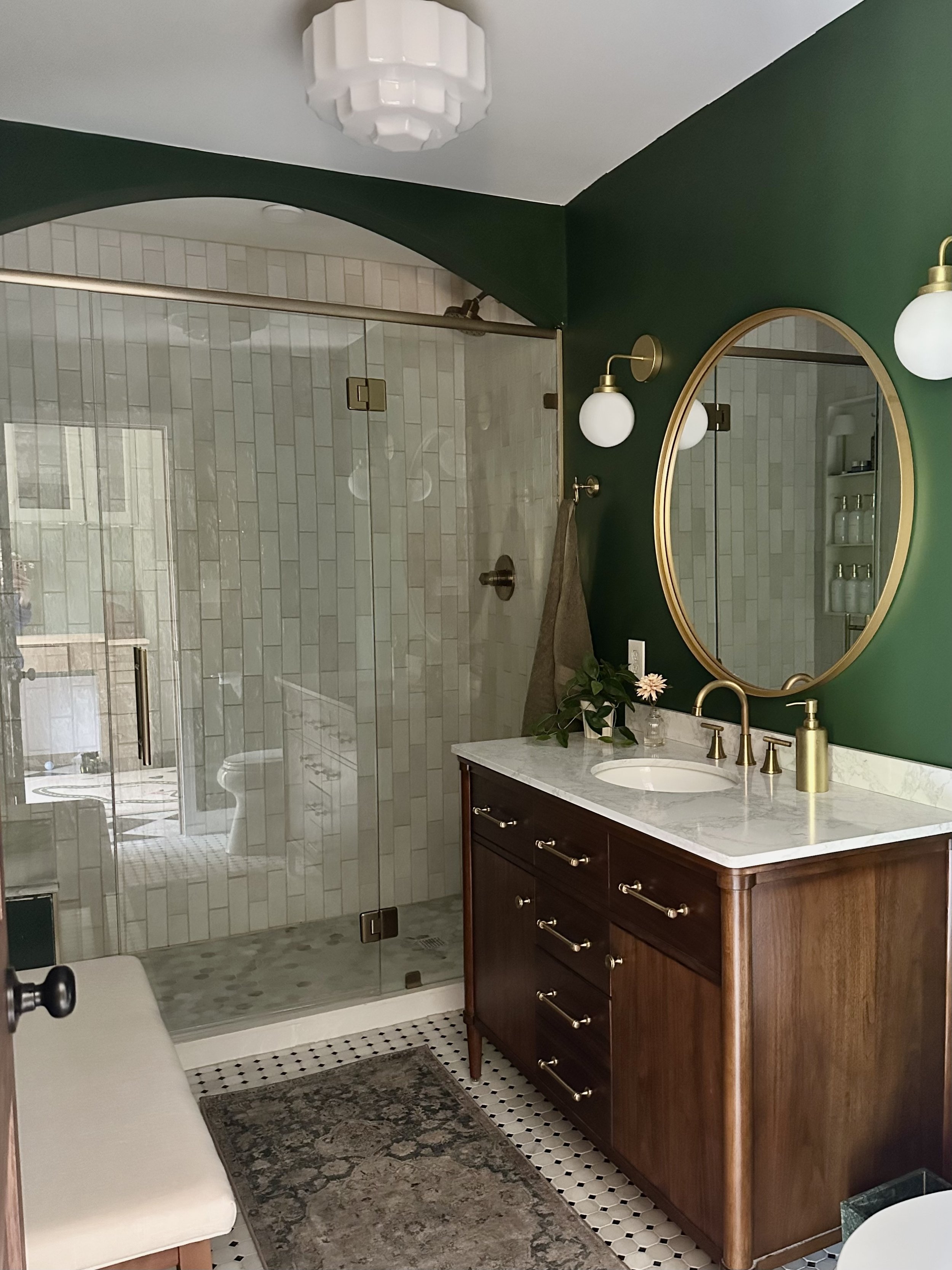 A bathroom with green painted walls, a wooden vanity with a marble countertop, a round mirror, and decorative lighting fixtures. The shower has glass doors and white tiled walls with a horizontal pattern.