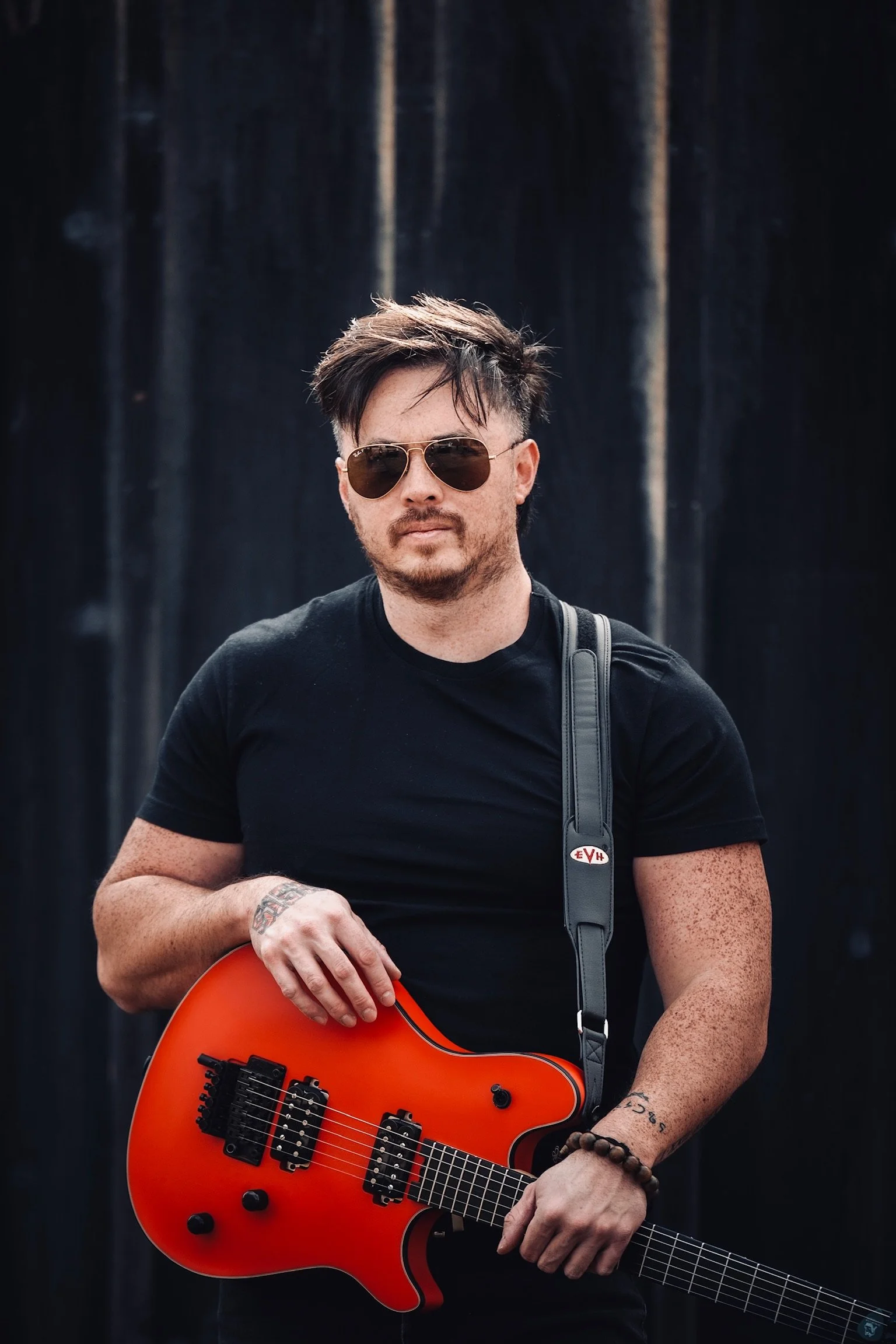 A man wearing sunglasses and a black t-shirt holding a red electric guitar against a dark background.