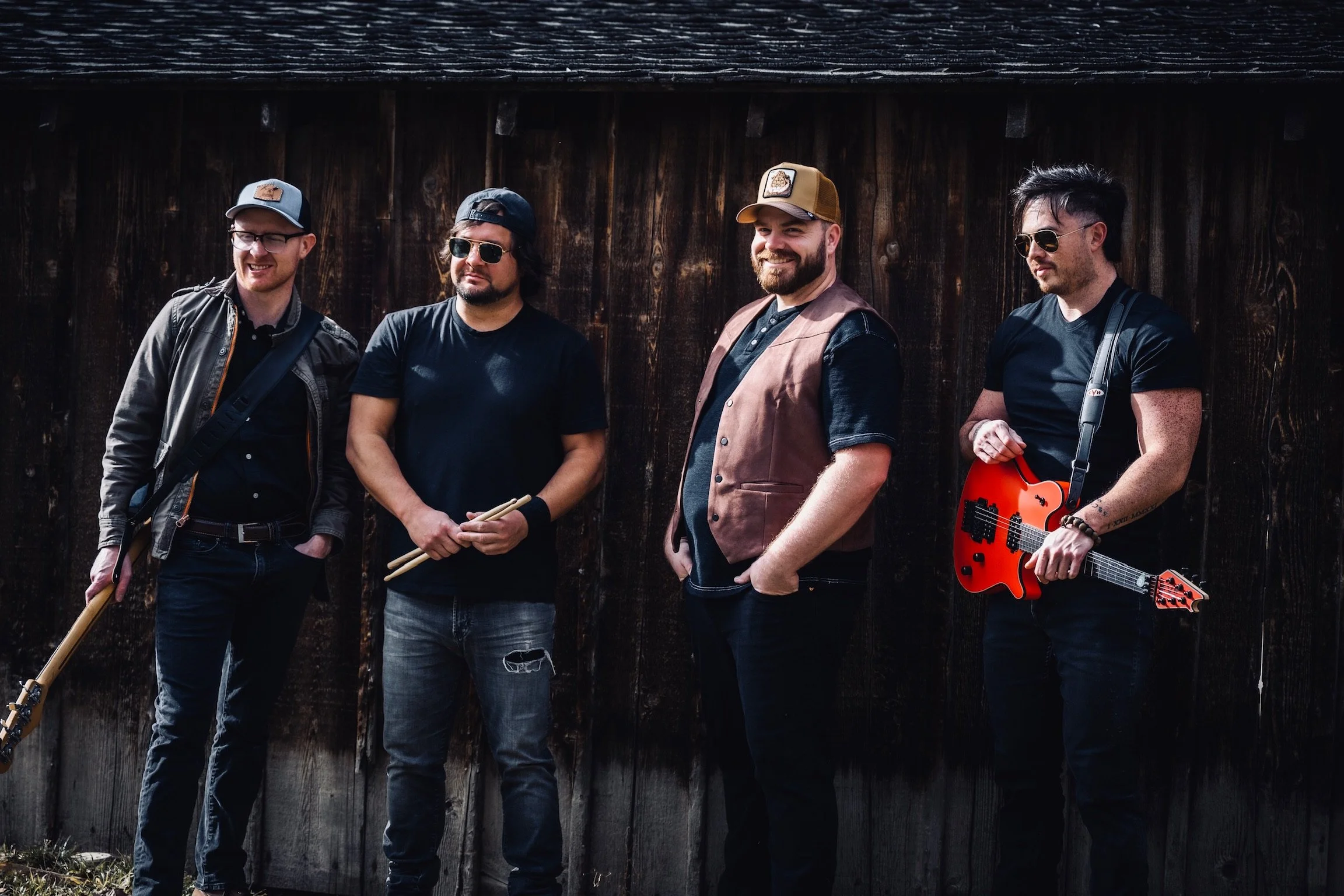 Four men standing outdoors against a dark wooden fence, holding musical instruments, dressed casually with sunglasses and hats, posing for a photo.