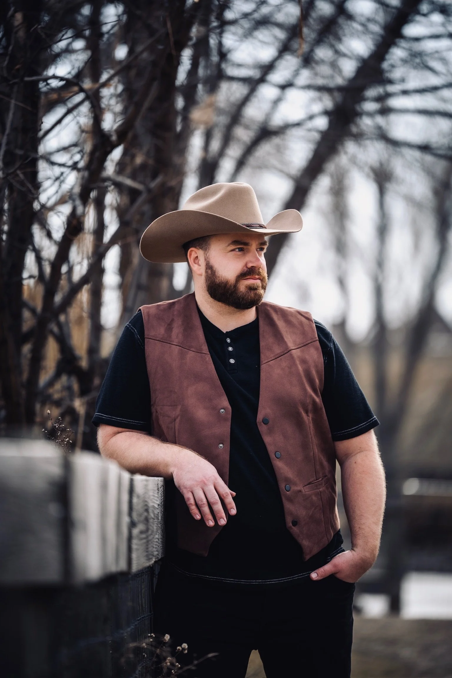 A man with a beard and a cowboy hat leaning on a wooden railing outdoors, with leafless trees in the background.