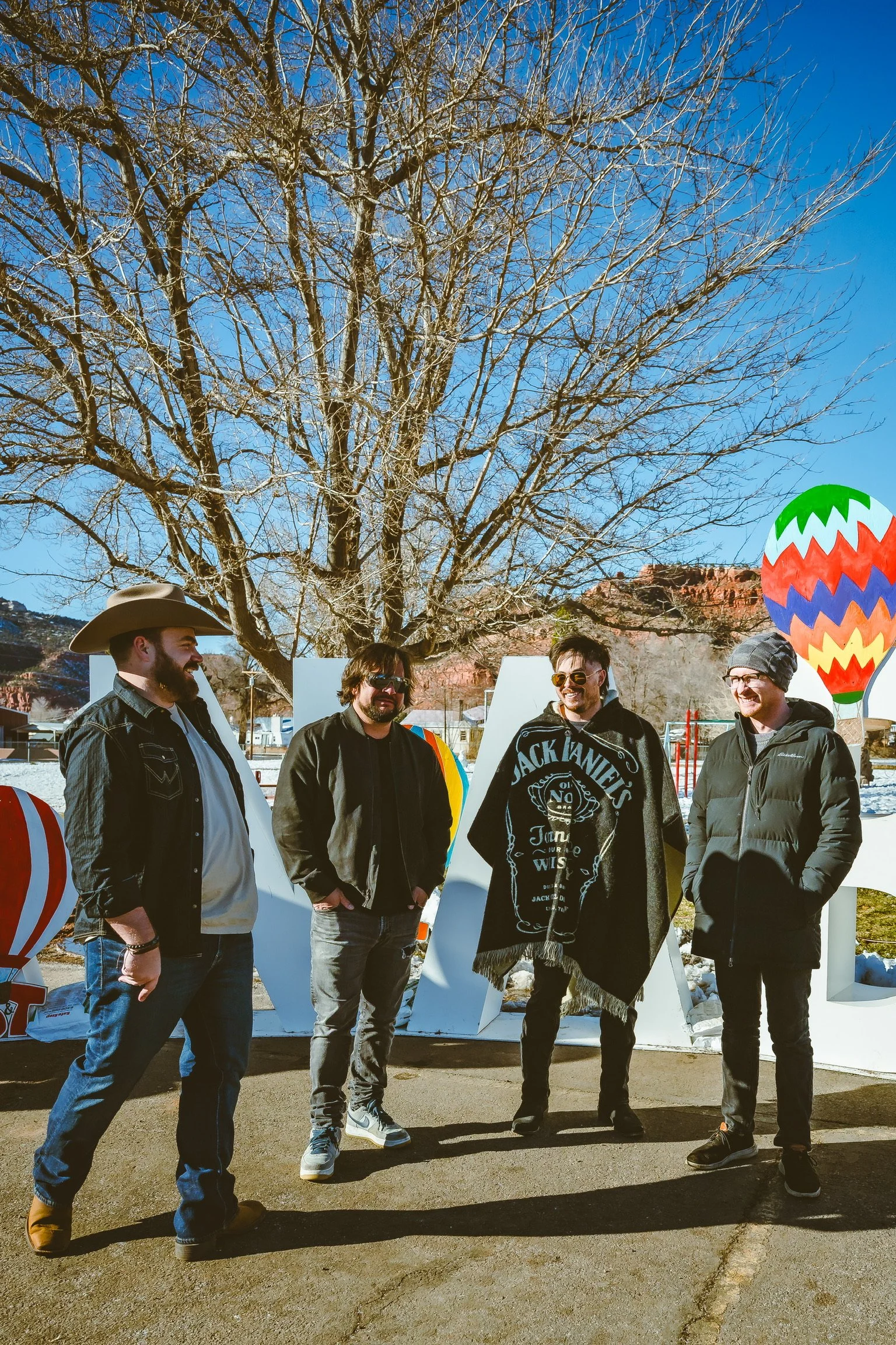 Four men stand outdoors near a large, leafless tree, smiling and talking. They are dressed in casual winter clothing, with one wearing a cowboy hat and another a Jack Daniel's blanket. Behind them are colorful decorative hot air balloons and large wh
