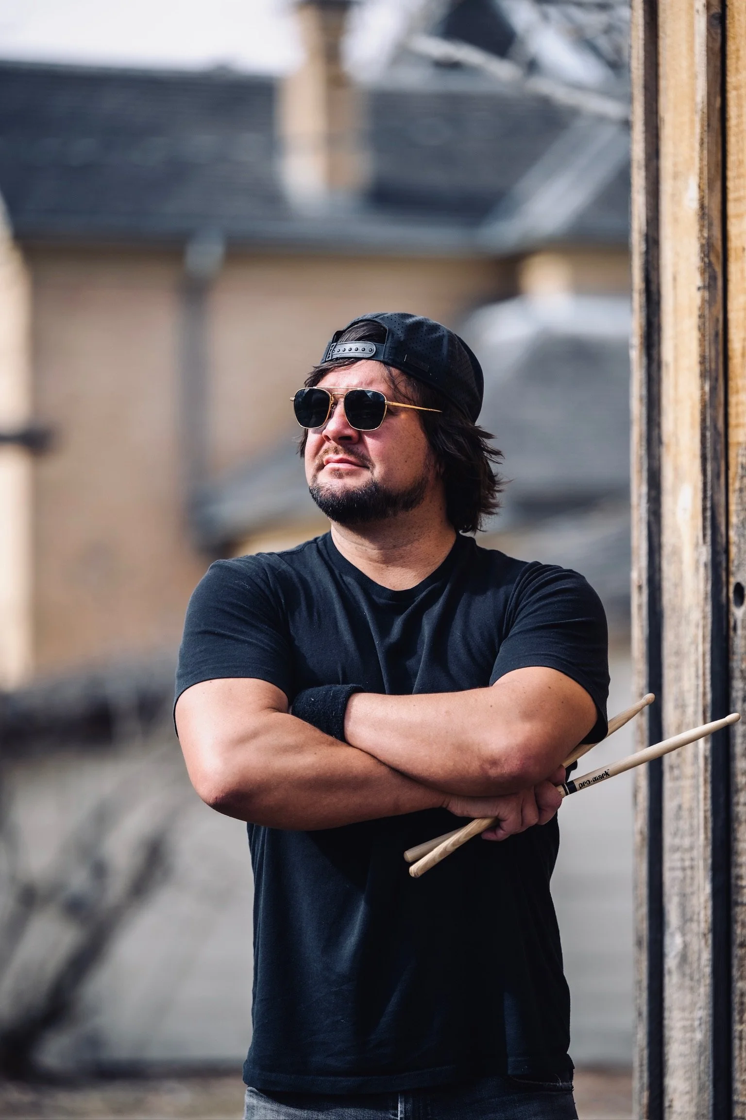 A man with dark hair, sunglasses, and a beard wearing a black cap and black t-shirt, standing outdoors with crossed arms and holding drumsticks, near a wooden structure.