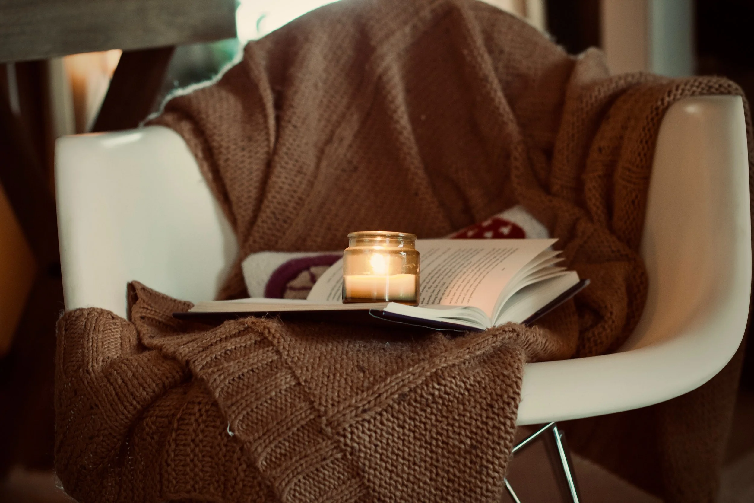 White chair with a brown knit blanket, an open book, and a small lit candle.