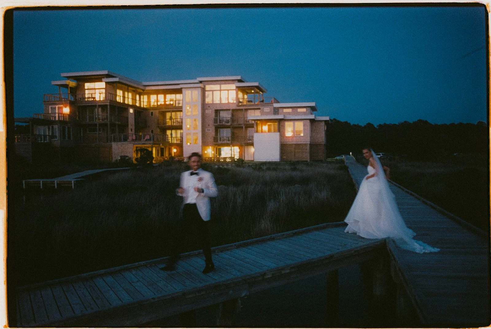 Bride and groom on the boardwalk at dusk, with the lit flagship house at Edgecamp behind them