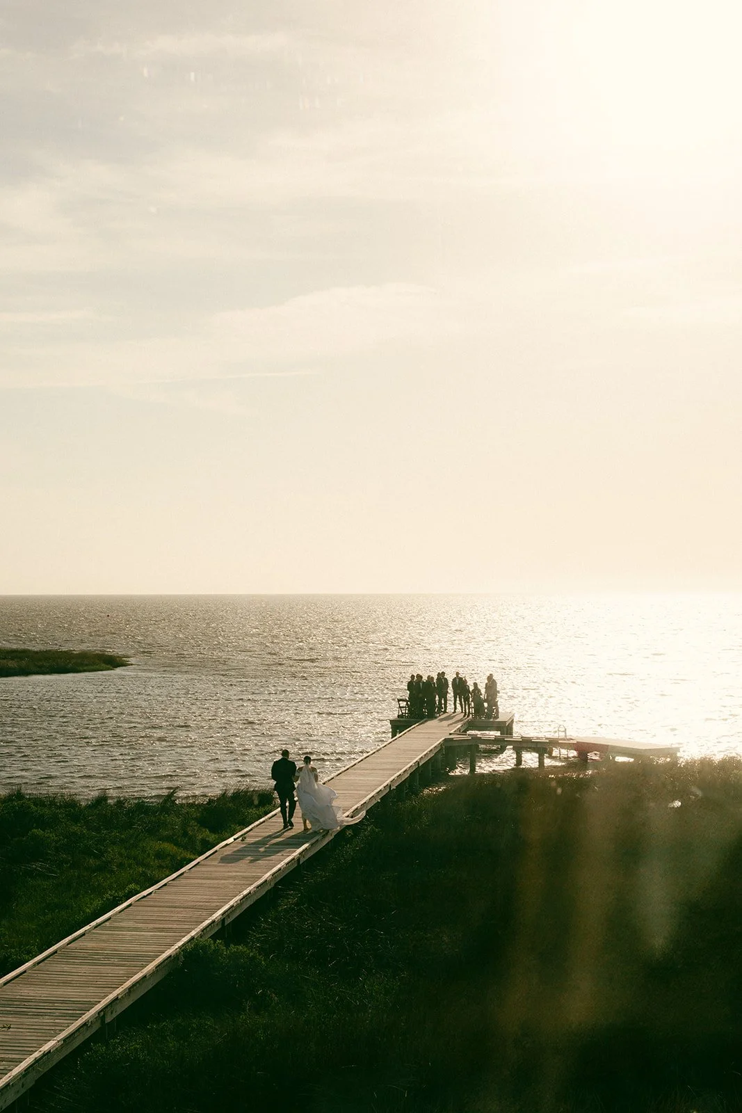 aCouple walking the boardwalk to a dock ceremony on the Pamlico Sound at sunset, guests waiting at the end