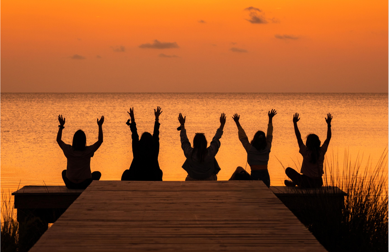 Five people sitting on a dock by the water with arms raised during sunset, with pink and orange sky and calm water