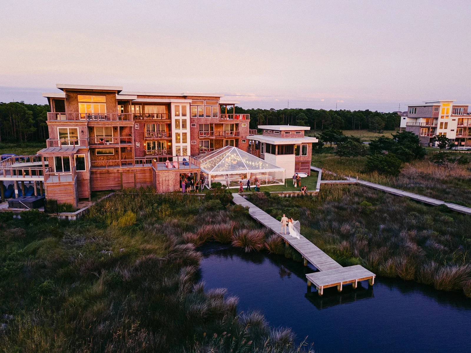 aSunset aerial of Edgecamp's flagship house and a lit clear-top reception tent, with a couple on the boardwalk