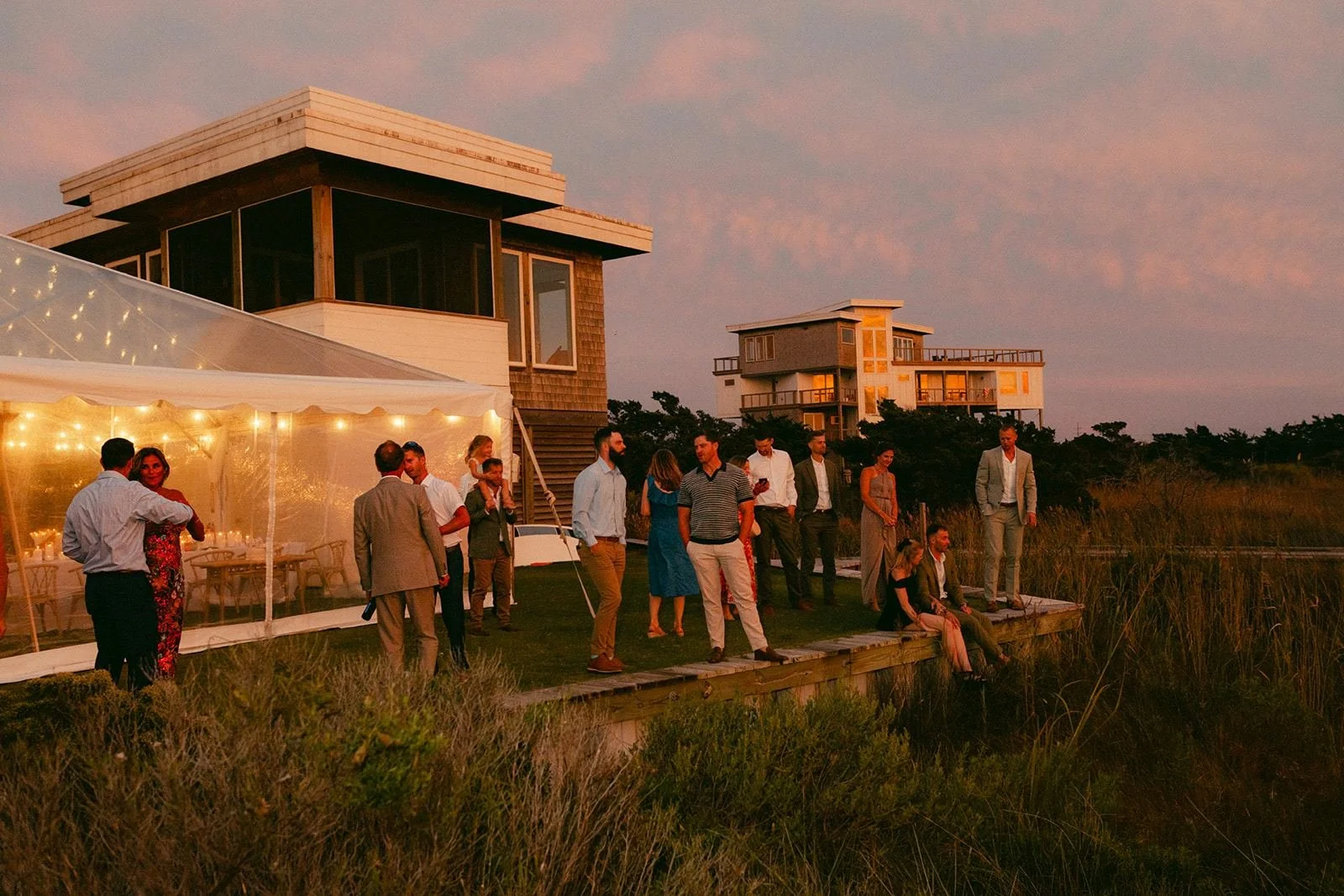 Wedding guests on the boardwalk at golden hour at Edgecamp, the tent lit beside the house.