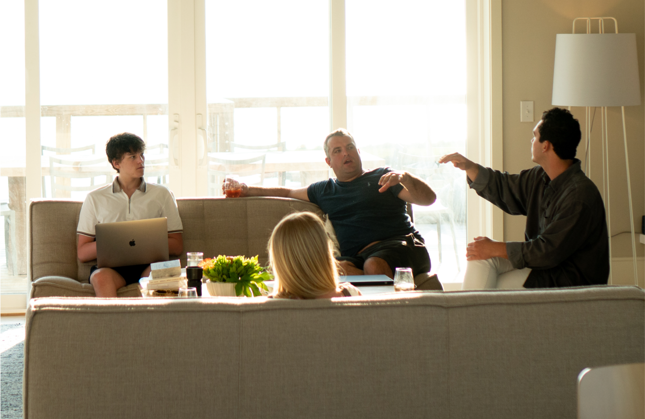 A group of four people having a discussion in a living room, with sunlight coming through large glass doors in the background. One person is sitting on a sofa with a laptop, another is leaning back with a drink, and two others are engaged in a conversation, one gesturing with their hand.