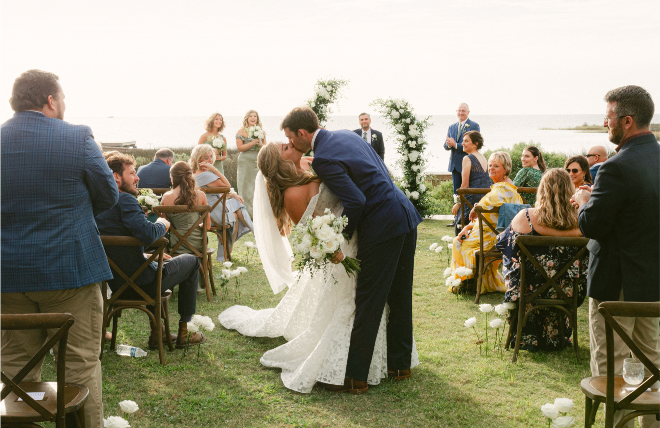 A newlywed couple sharing a kiss during their outdoor wedding ceremony by the water, with guests seated on either side, floral arrangements, and a romantic arch in the background.