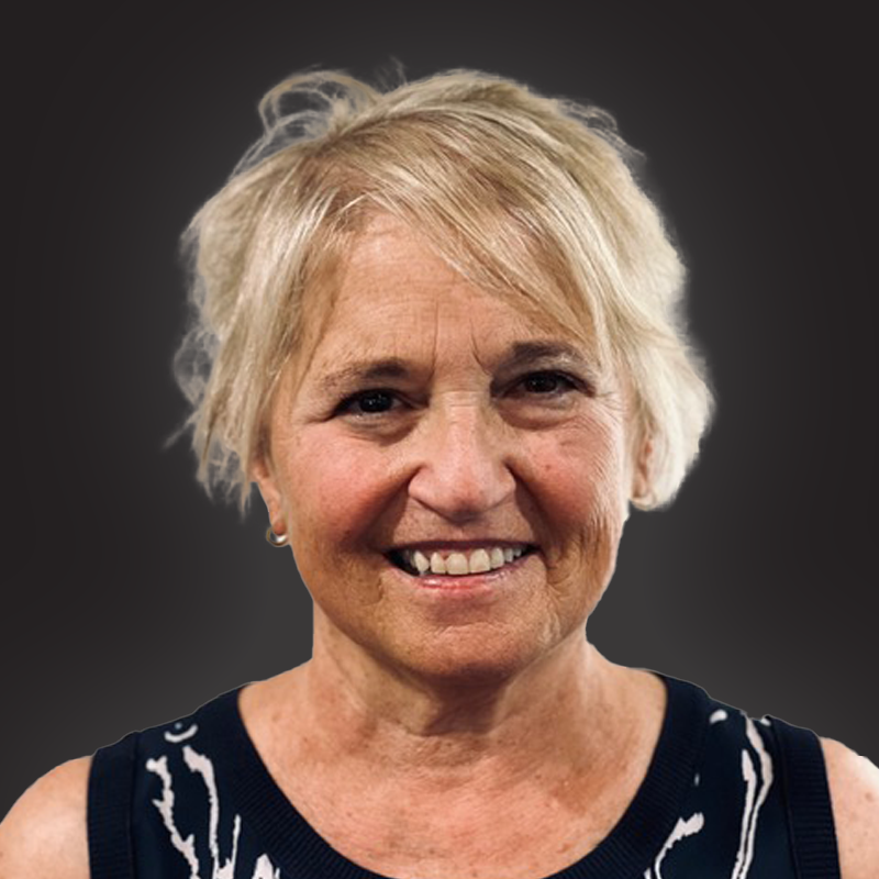 A smiling middle-aged woman with short blonde hair wearing a black sleeveless top with white patterns, standing against a dark background.