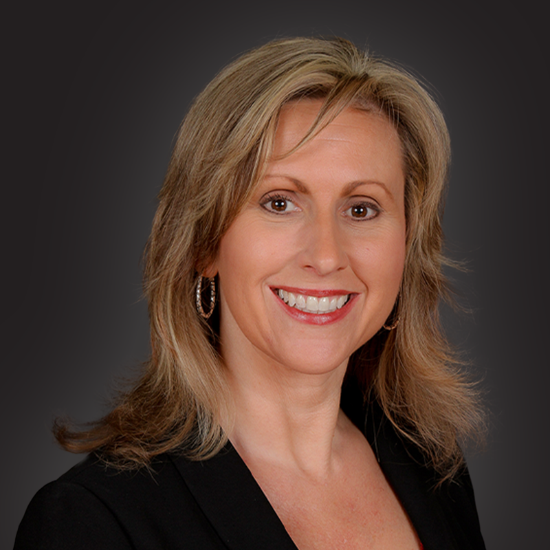A professional woman with blonde hair smiling, wearing a black blazer and hoop earrings, against a dark background.