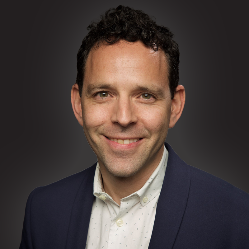 Headshot of a smiling man with dark, curly hair wearing a white collared shirt and navy blazer against a dark background.