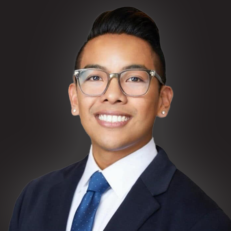 Portrait of a young man with glasses, wearing a suit and tie, smiling against a dark background.