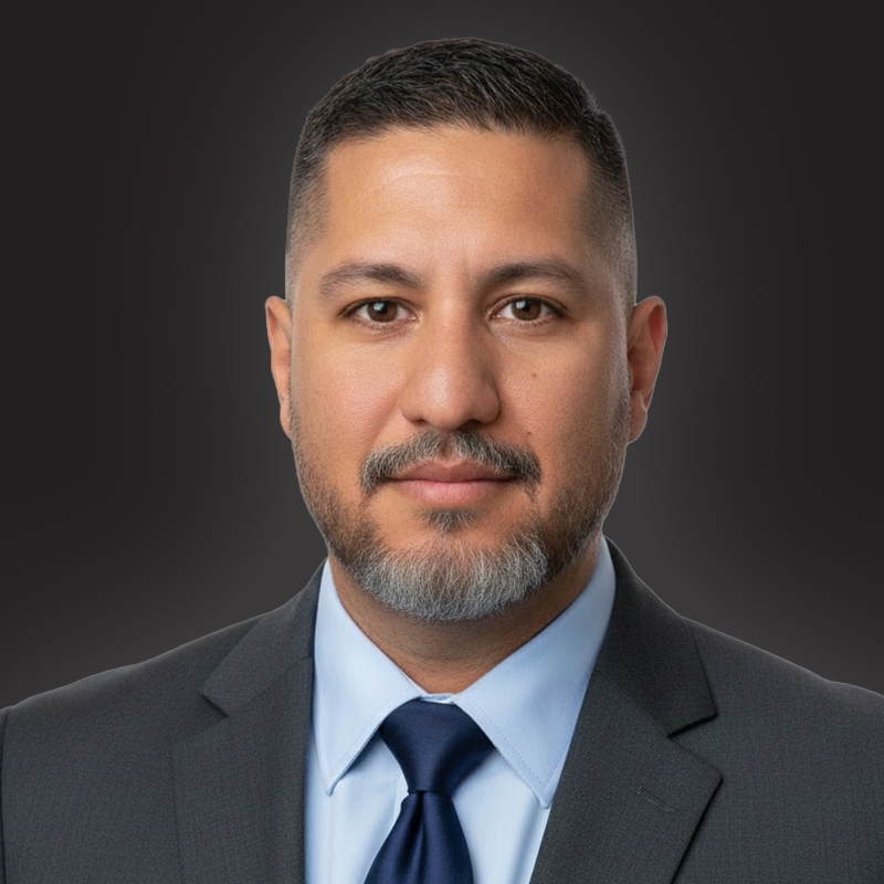 A portrait of a man in a business suit with short dark hair, beard, wearing a light blue shirt and dark tie, against a dark background.
