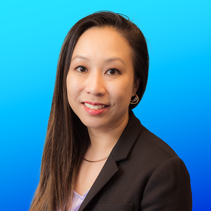 Portrait of a woman in a black blazer with long hair, smiling, against a blue gradient background.